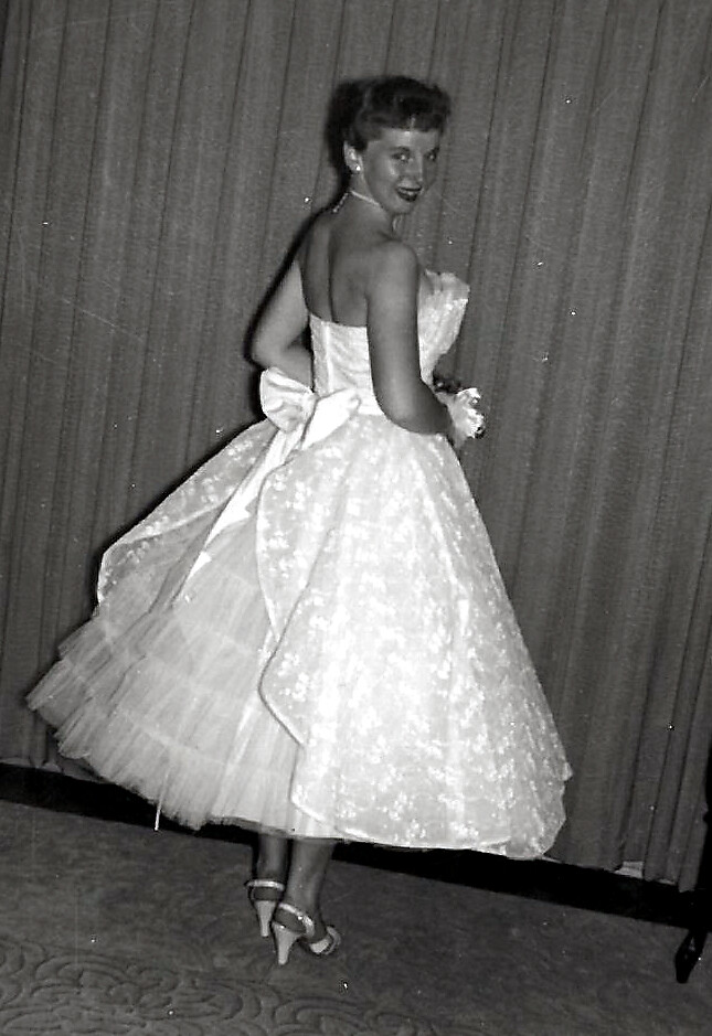 Back view of a woman in a strapless, full-skirted ball gown with a large bow at the waist, posing on a stage with curtains.