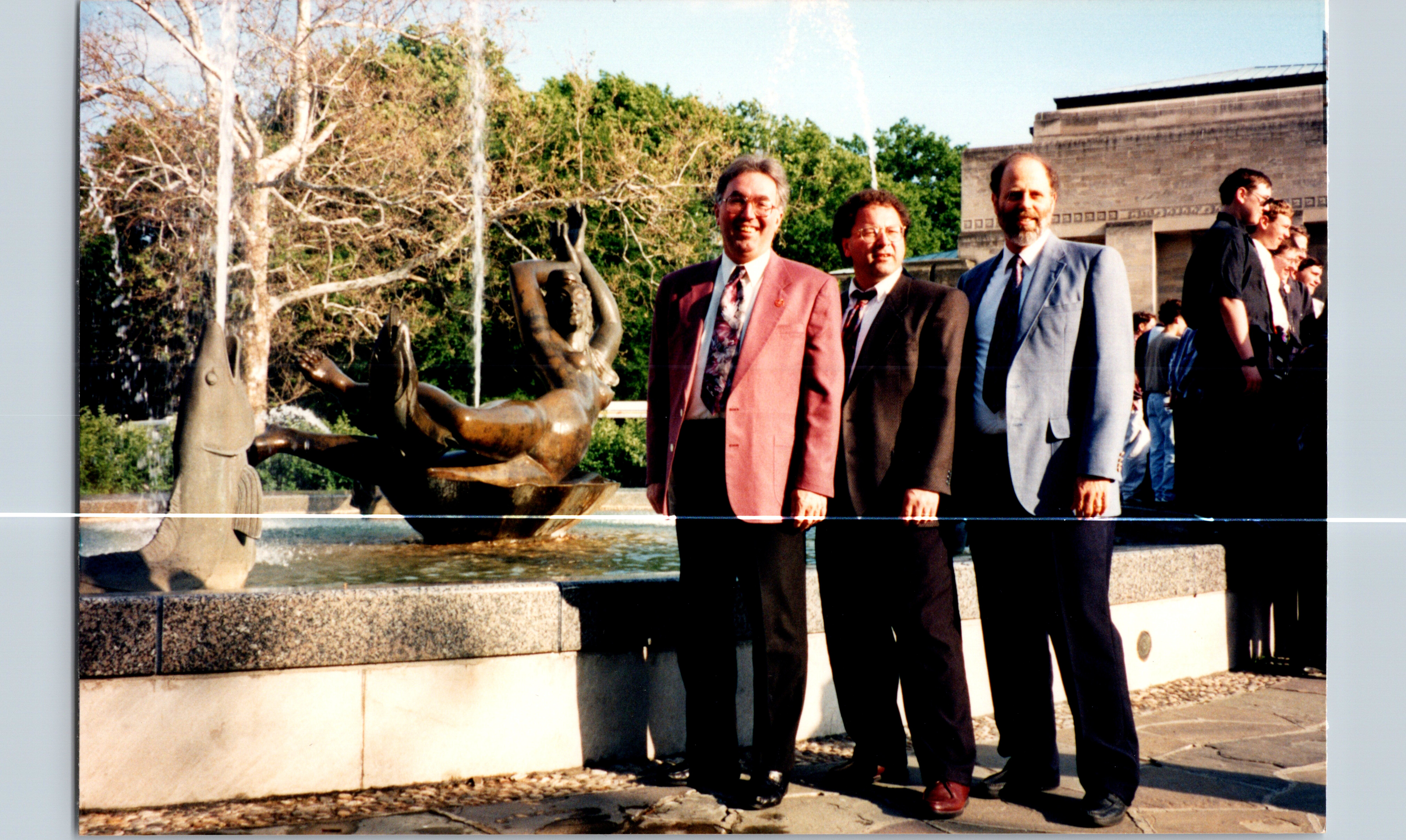 Group of men in business suits posing by a fountain statue outdoors, with trees and a building in the background.