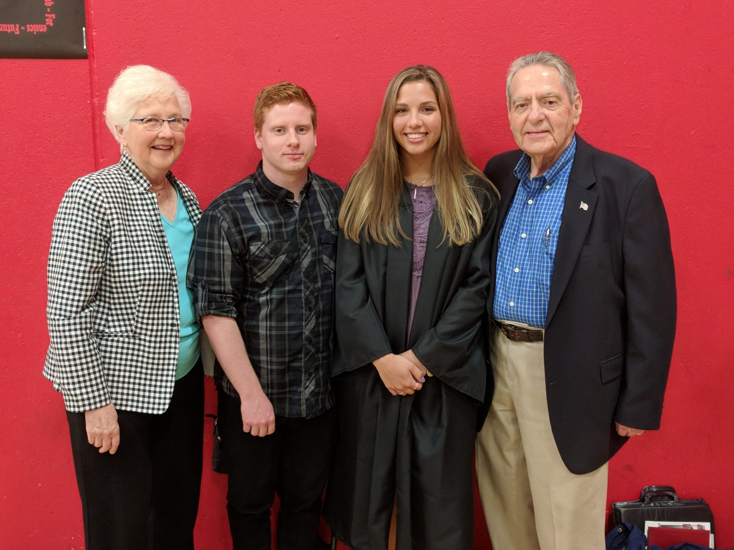 Four people posing for a photo in front of a red wall: an older woman, a young man, a smiling graduate in a black gown, and an older man in a blazer.