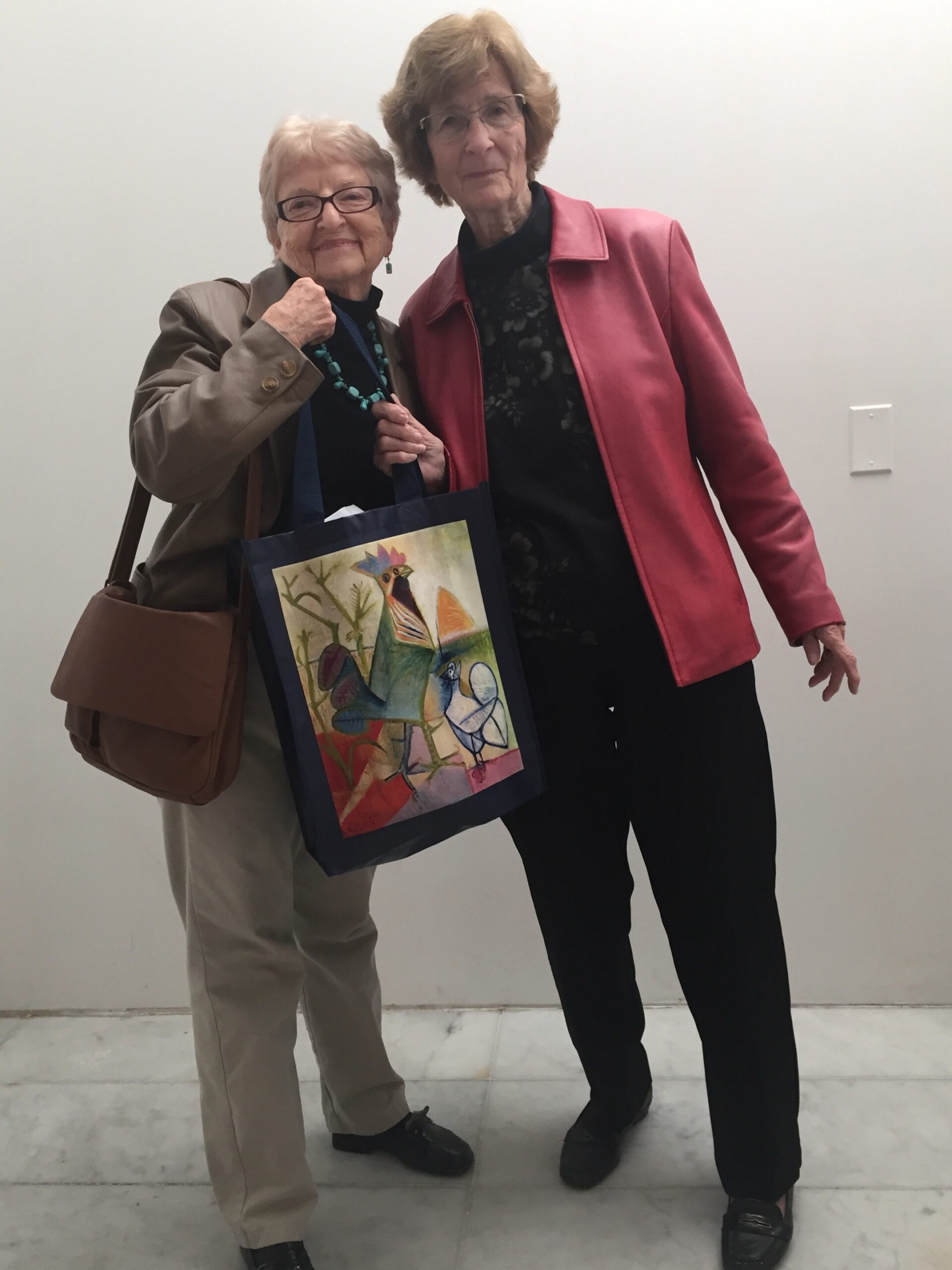 Two smiling elderly women pose together indoors; one wears a tan jacket and turquoise necklace, holding a colorful art tote bag beside her friend in a pink jacket.