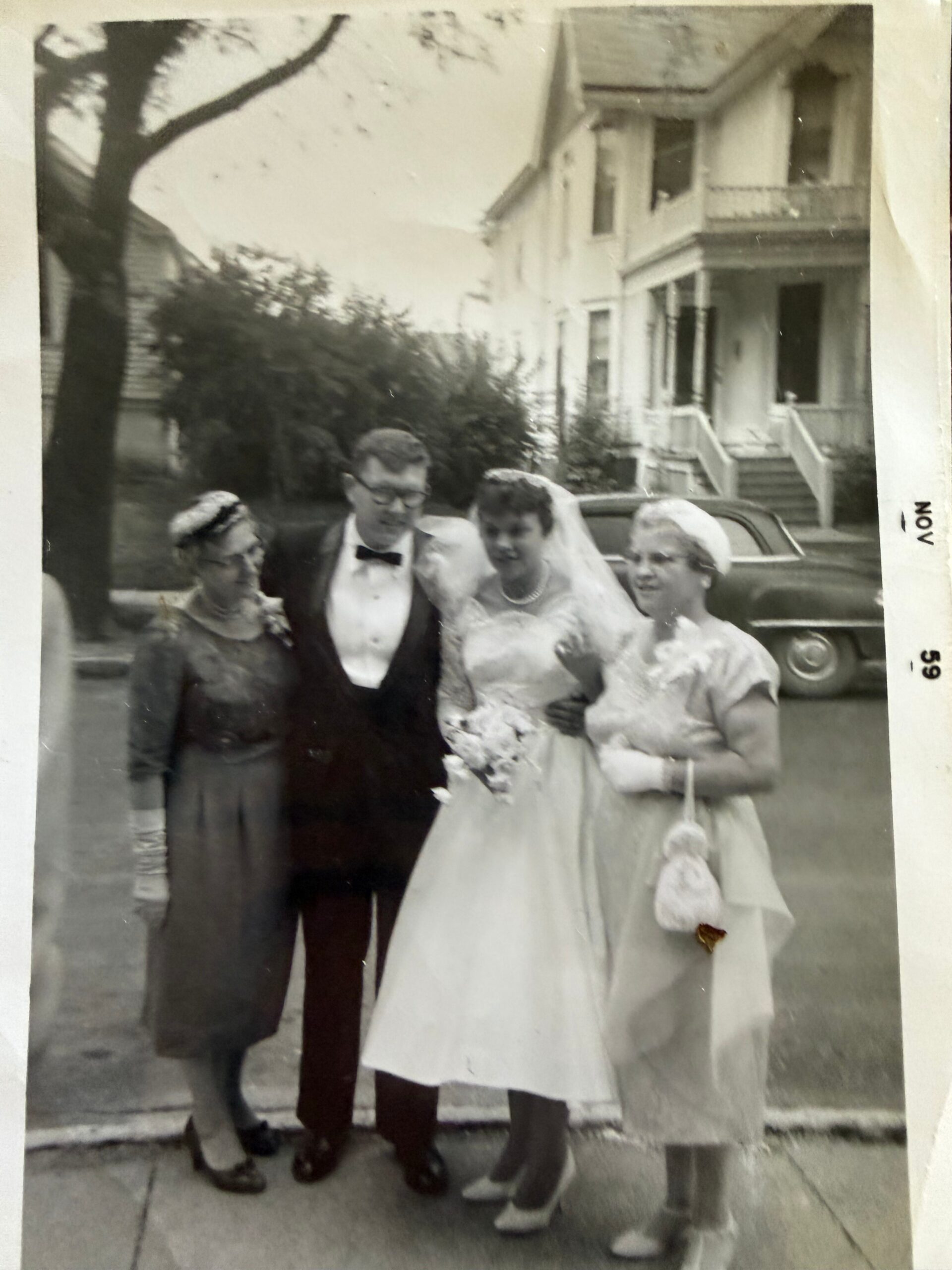Bride in white gown and veil flanked by groom in tuxedo and two guests outside a row of houses, posing for a wedding photo.