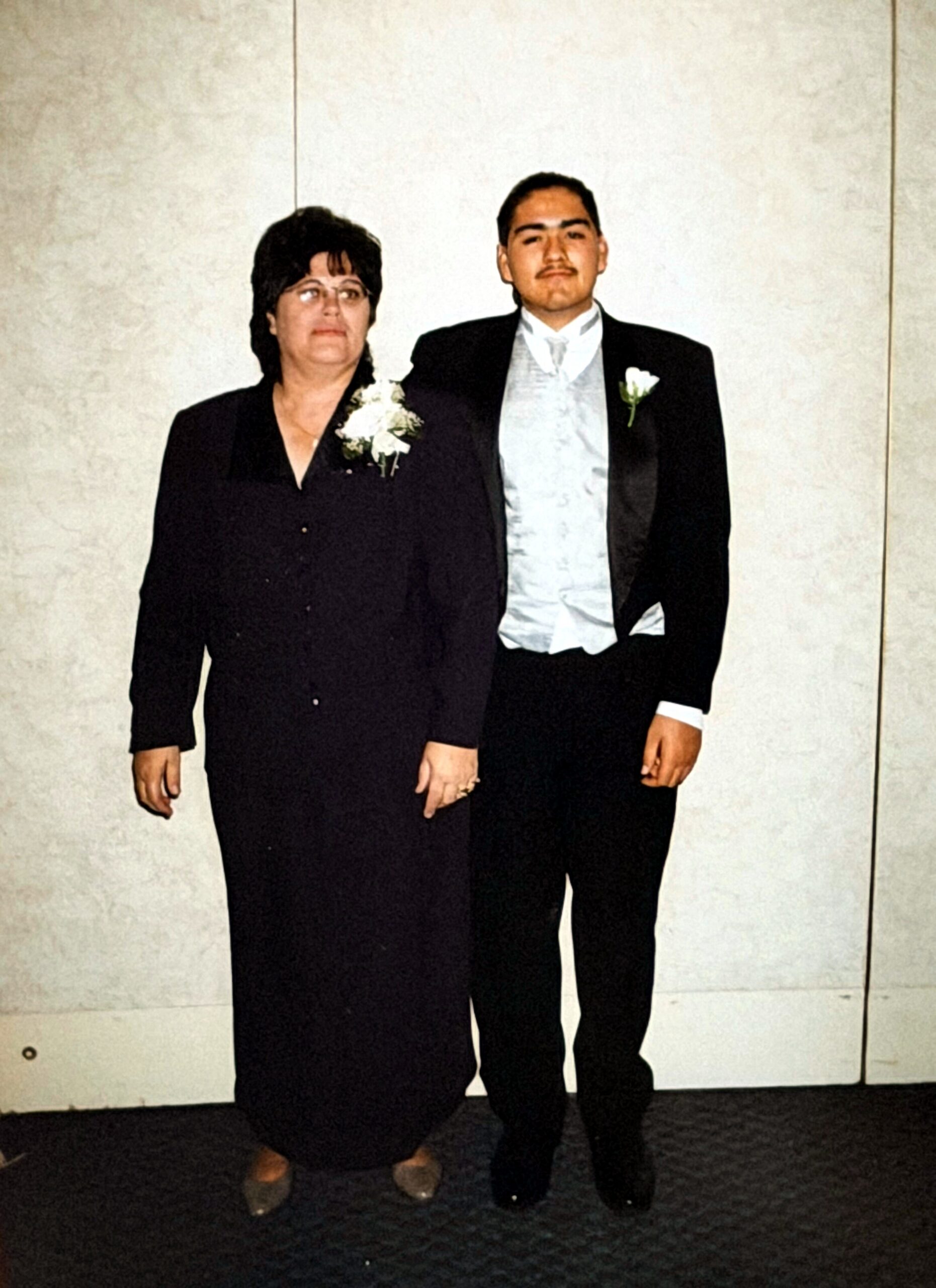 Woman in a black dress with a white corsage stands beside a man in a tuxedo with a boutonniere, posing for a formal photo against a light wall.
