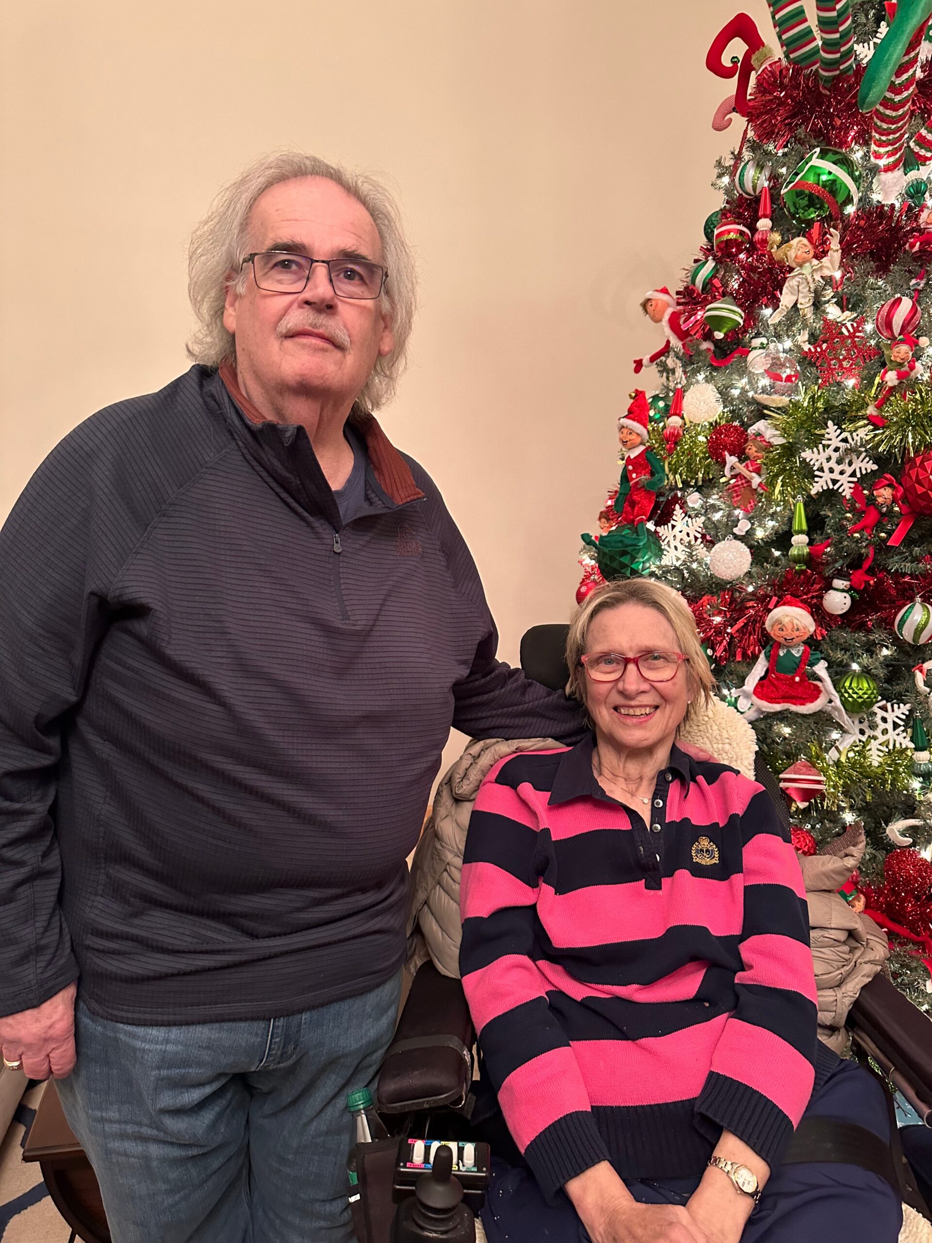 Older couple posing indoors beside a festively decorated Christmas tree with red and green ornaments and tinsel.