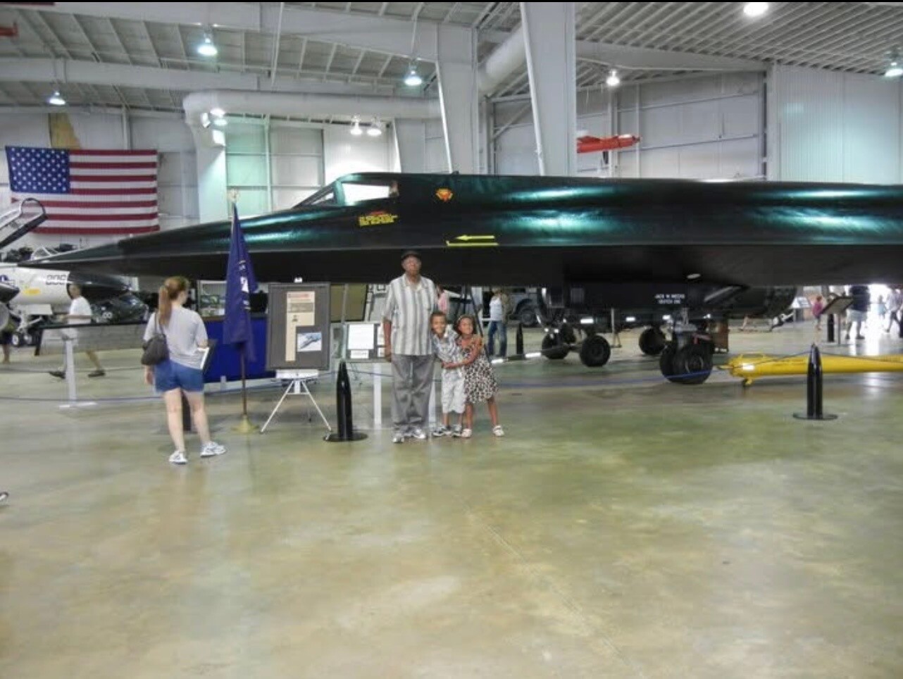 Indoor aircraft hangar with a large dark jet on display; a family poses in front while visitors walk around and an American flag hangs in the background.