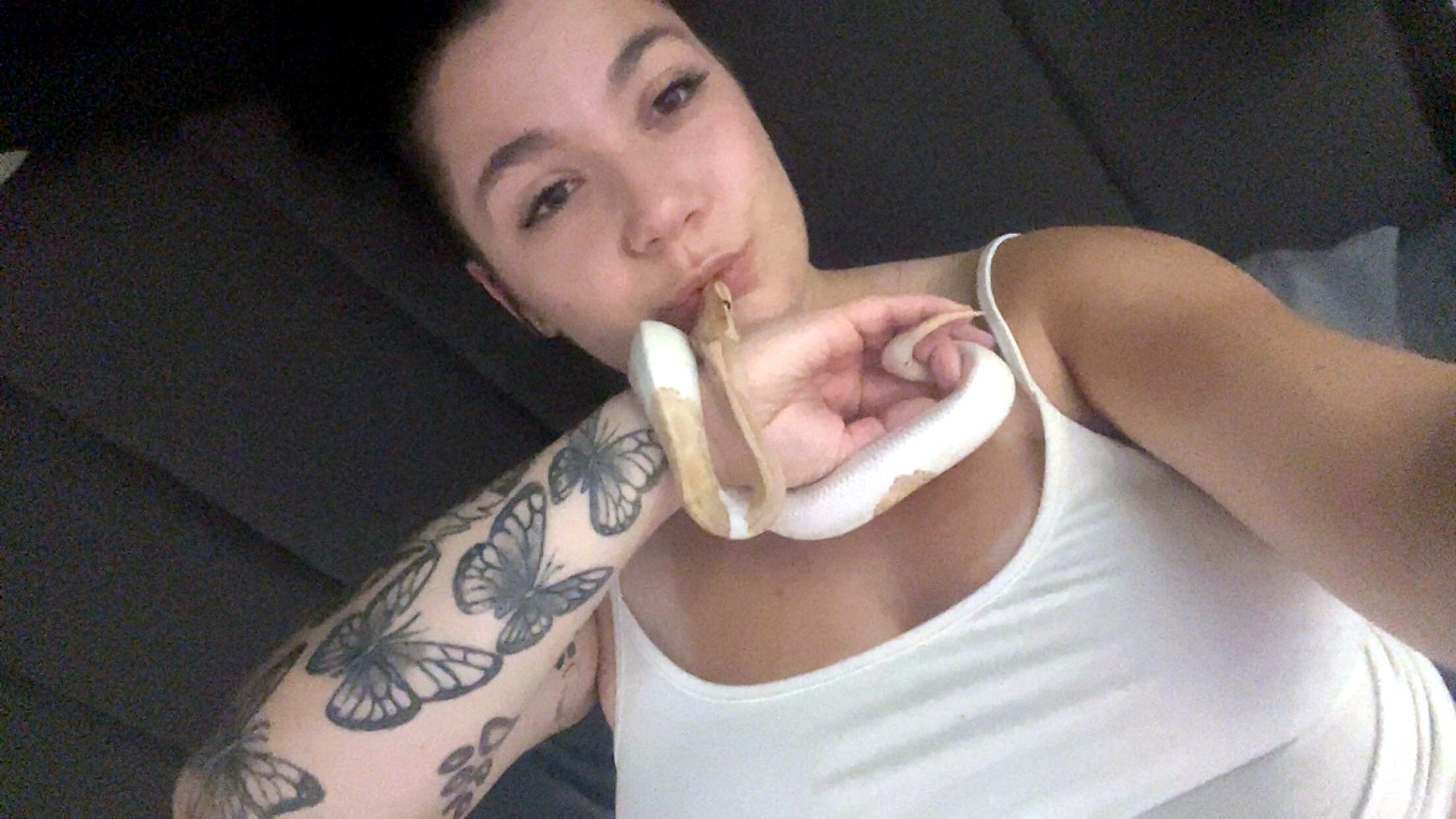 Woman lying on her side with a light-colored snake draped around her neck and shoulders, displaying butterfly tattoos on her arm.