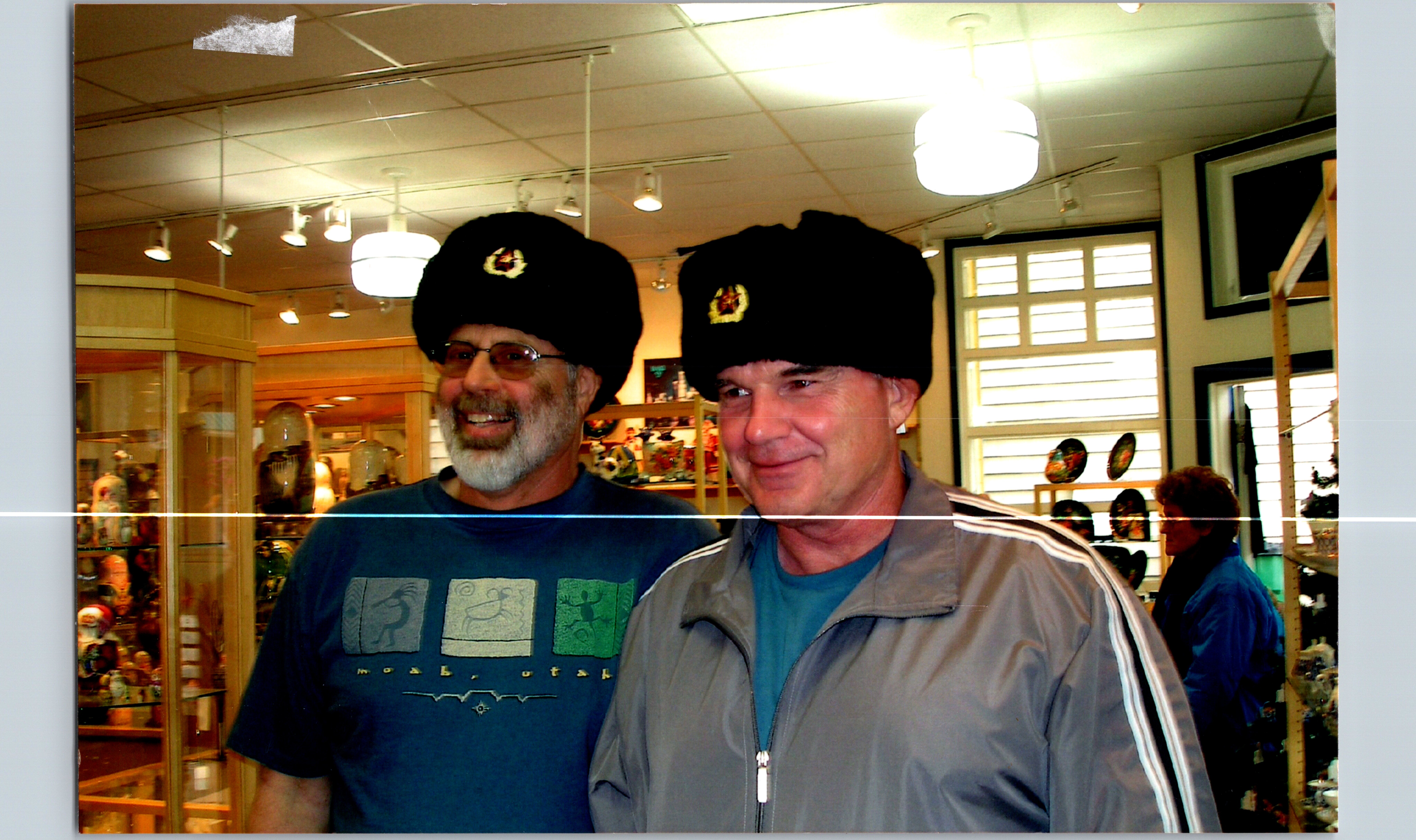 Two men in a store pose for a photo, both wearing large black fur hats with emblems, smiling at the camera.