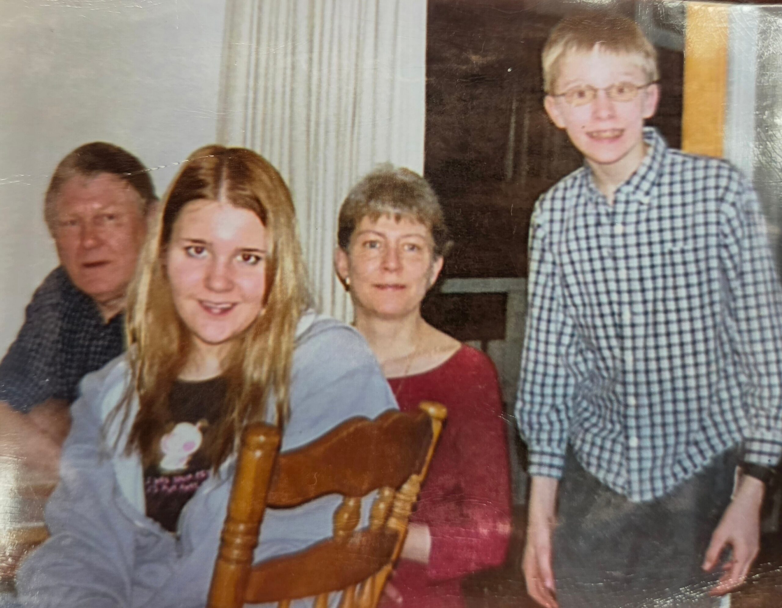 Family group indoors posing for a photo: a teen girl in gray sweatshirt sits in front, adults nearby, and a tall boy in a checkered shirt stands on the right.
