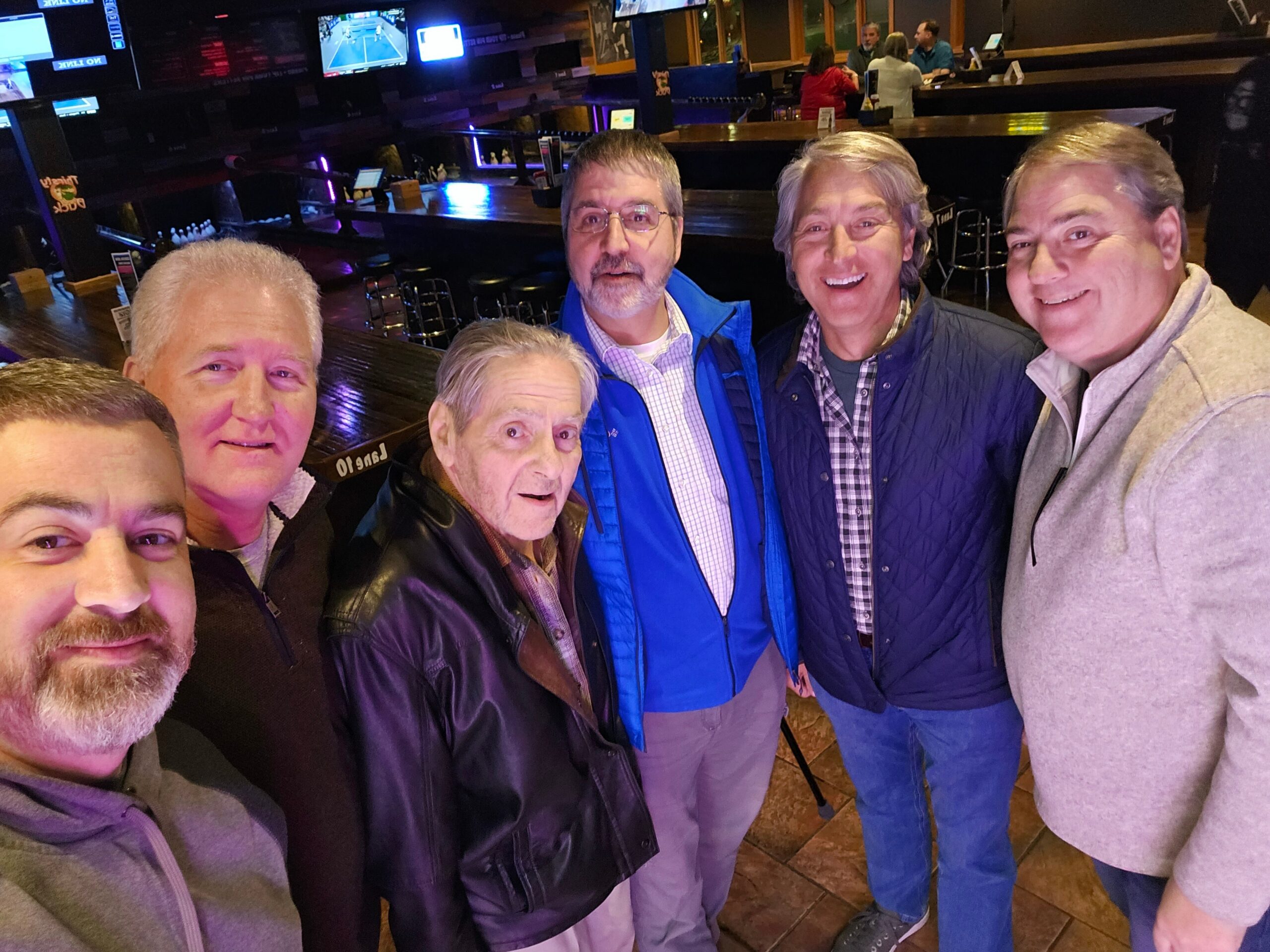 Five men in casual jackets take a group selfie at a dimly lit bar, smiling with a counter and screens behind them.