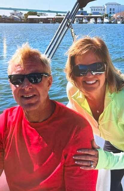 Smiling couple wearing sunglasses on a boat by the water with a marina in the background.