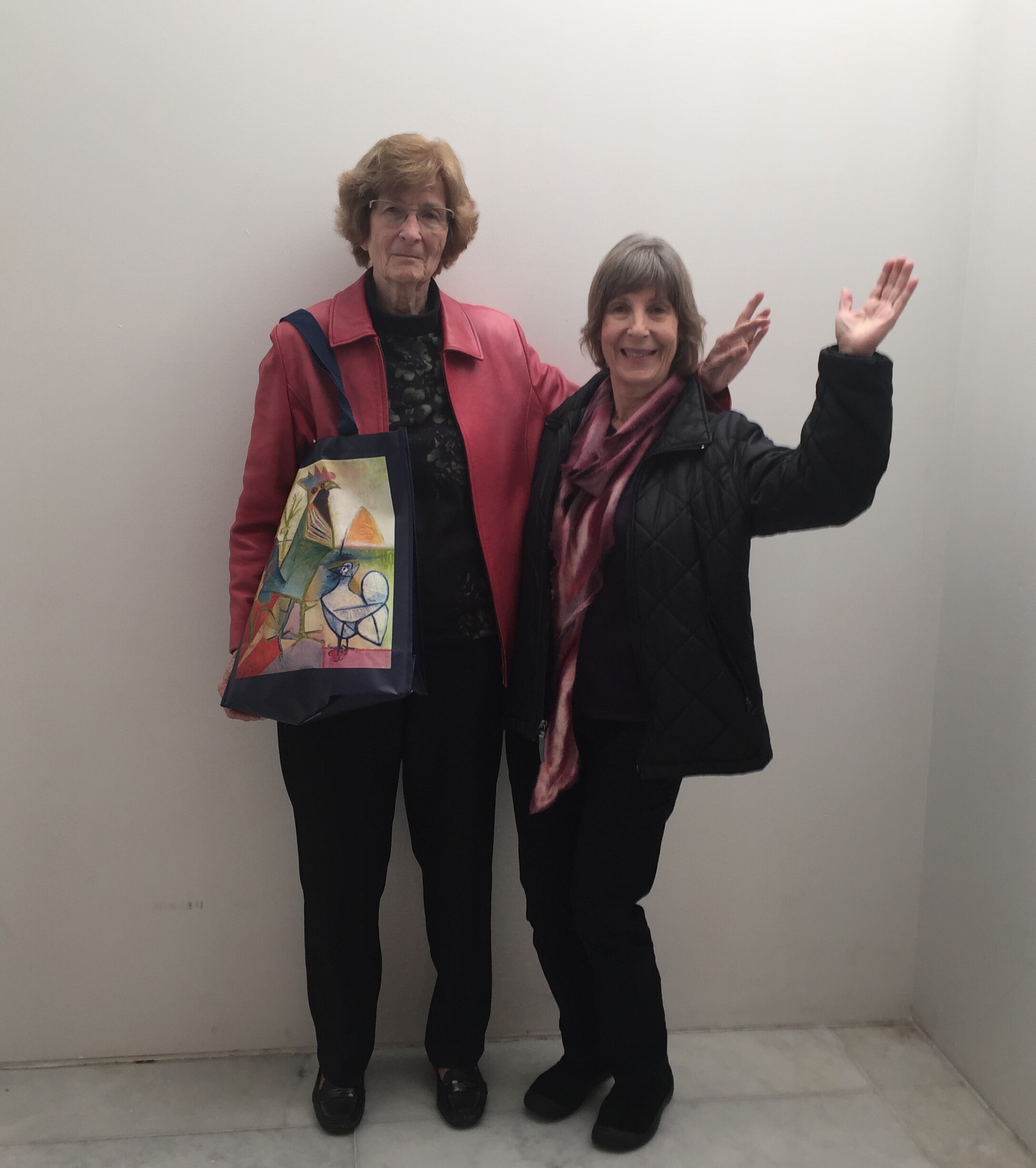 Two older women pose in a plain hallway; left wears a red jacket with a colorful tote bag, right wears a black jacket and raises a hand, smiling.