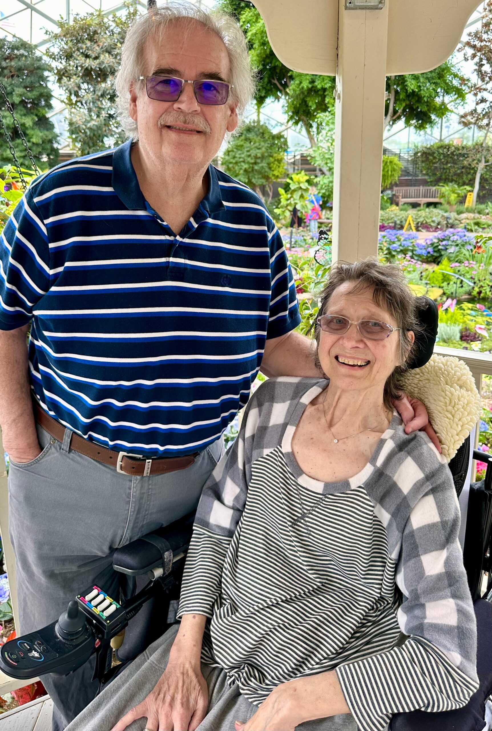 Smiling elderly man in a striped polo stands beside a seated woman in a wheelchair in a colorful garden behind a pergola. Placeholder device at wheelchair armrest is visible.