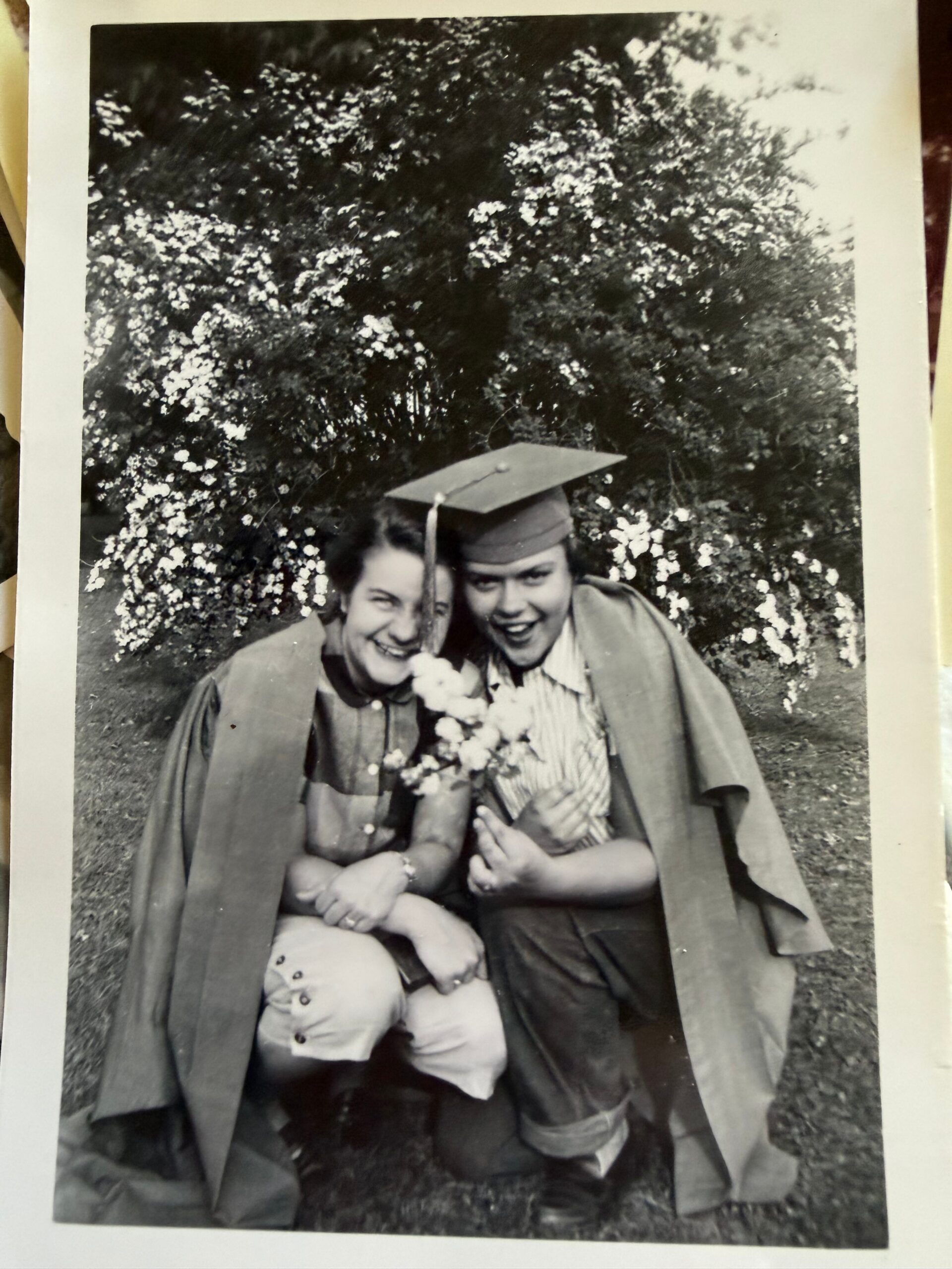 Two graduates in caps and gowns pose outdoors in front of blooming bushes; one holds a bouquet.