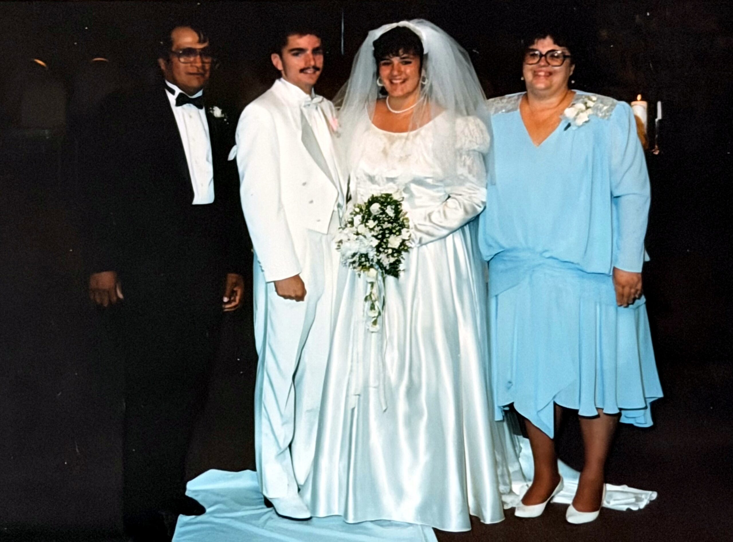 Bride in a white gown with veil holding a bouquet, standing with groom in a white suit and two guests in formal attire at a wedding.