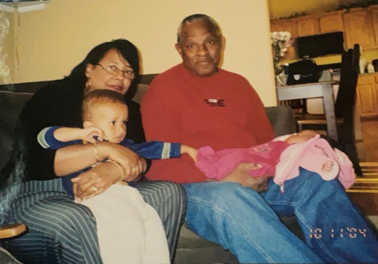 A multi-generational family portrait on a couch: a woman with glasses holding a toddler, beside an older man in a red shirt cradling a baby wrapped in pink blankets.