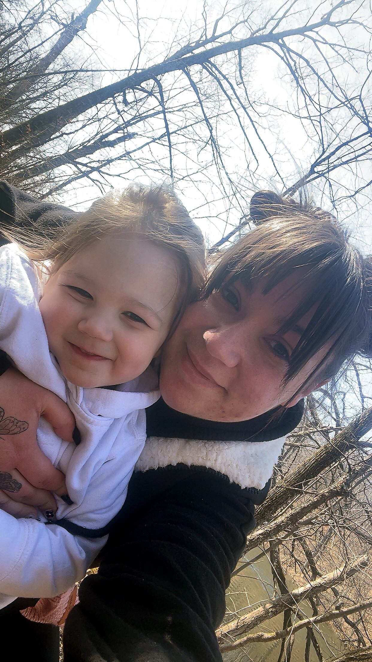 Smiling woman and young child in a close selfie outdoors on a sunny winter day, with bare tree branches in the background.