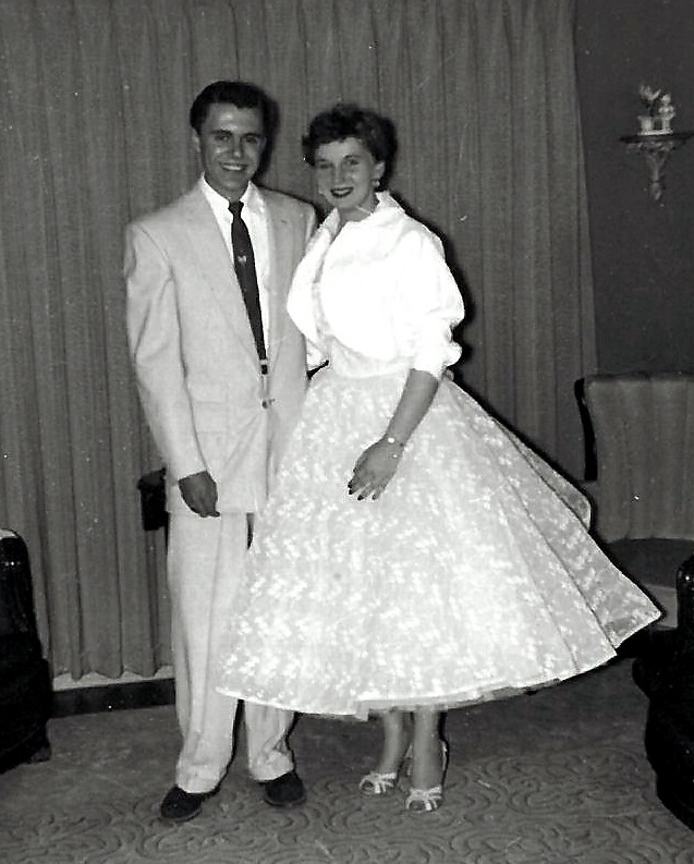 Couple in formal 1950s attire posing indoors; man in a light suit, woman in a white, full-skirted dress and heels, smiling for the camera.