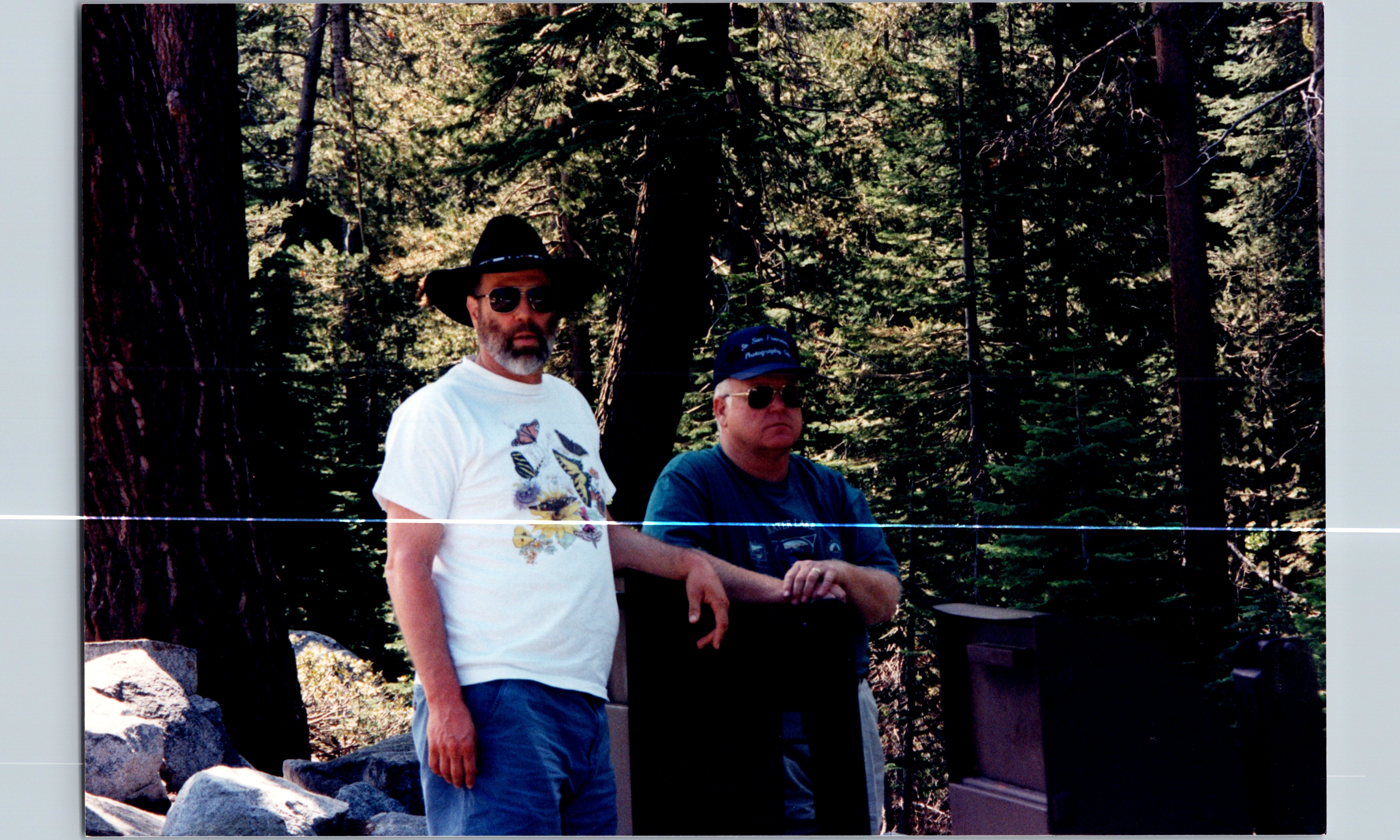 Two men wearing sunglasses and hats pose in a forested area with rocks nearby