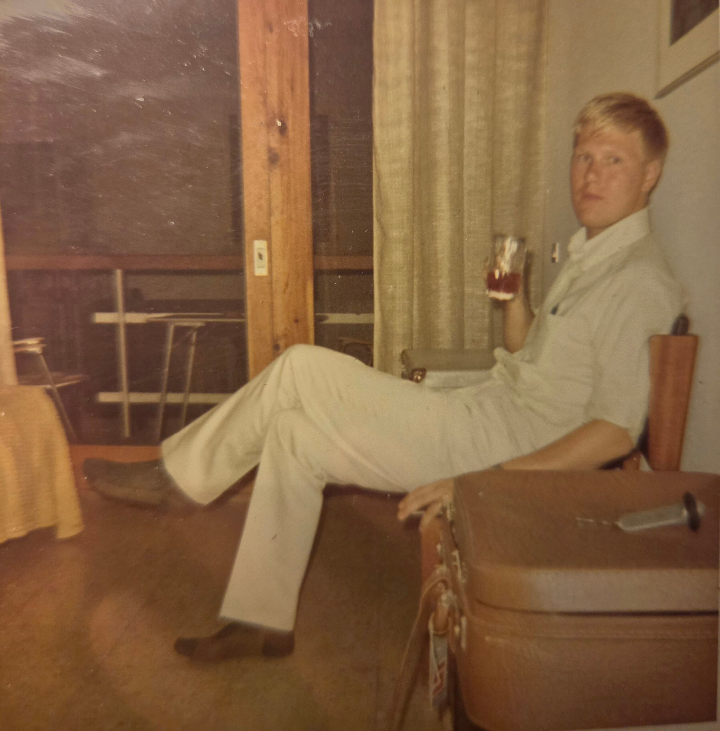 Portrait of a man with blond hair in a light shirt, seated indoors beside a wooden pillar and beige striped curtains.