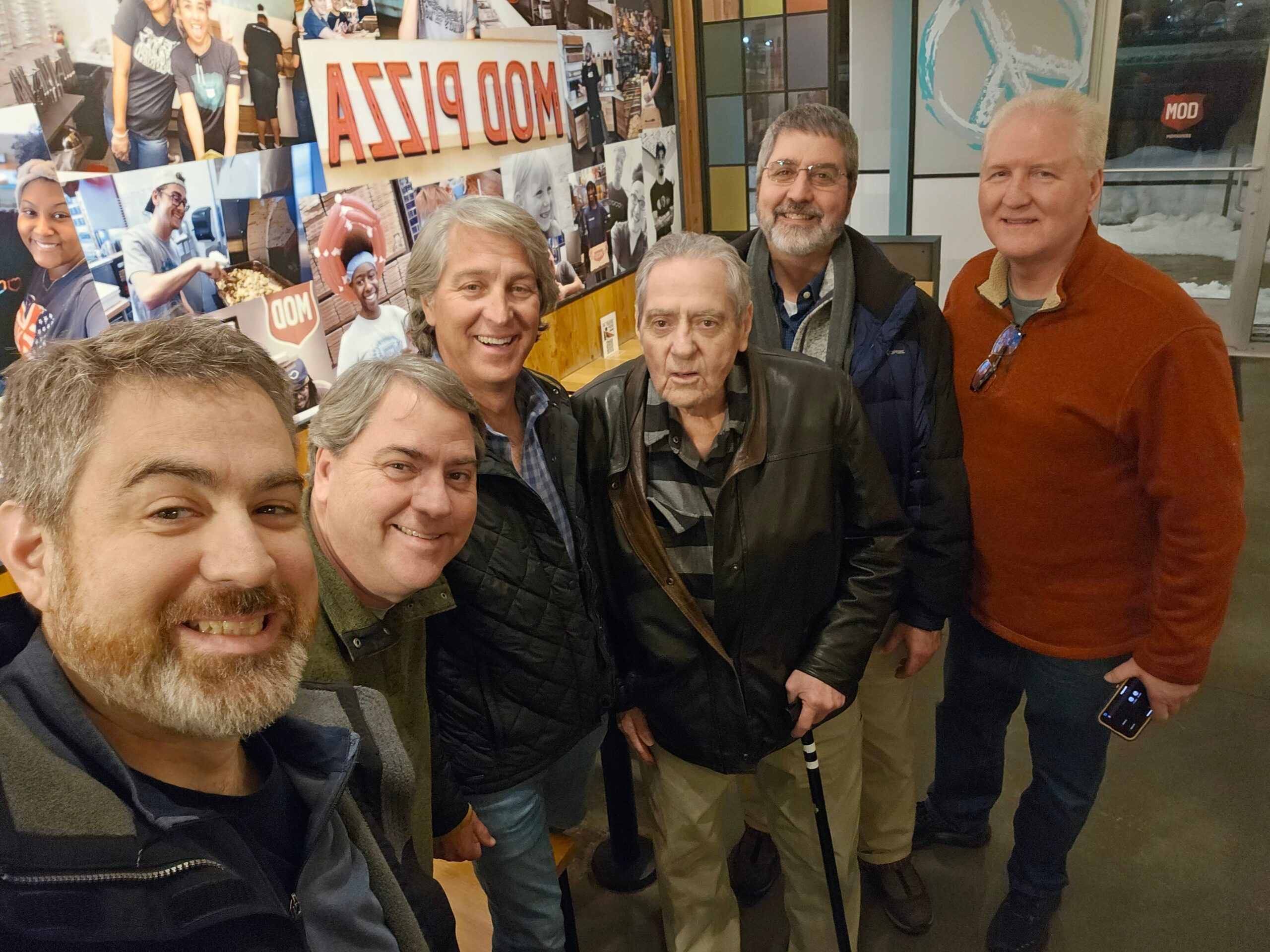 Group of men taking a selfie in a building lobby with a photo collage wall behind them.