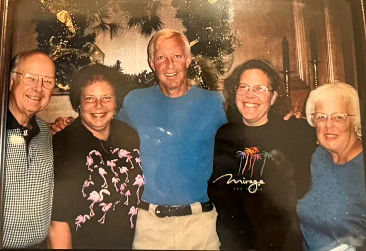 Five adults standing together indoors, smiling for a group photo with a Christmas tree in the background.