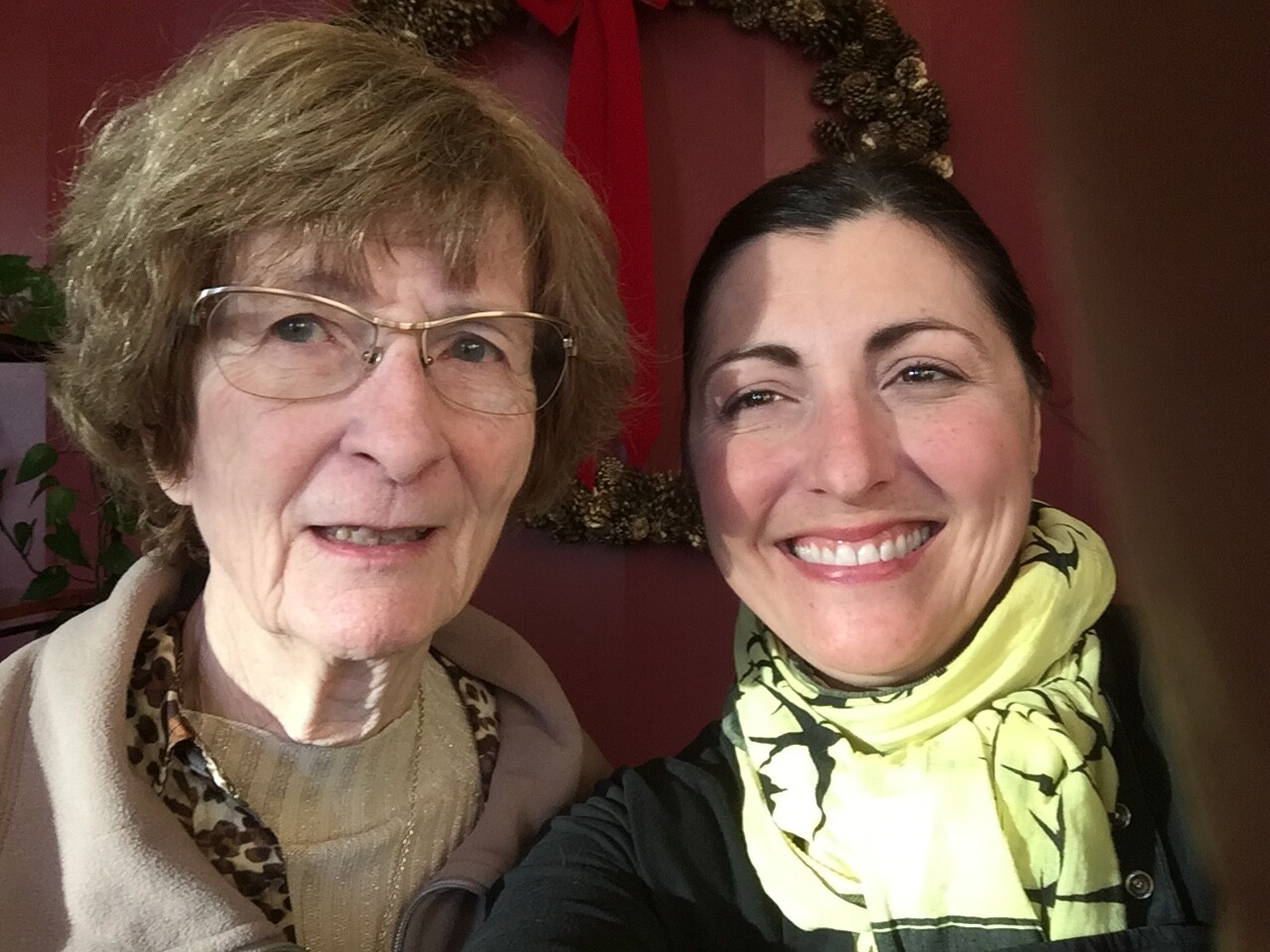 Two women posing for a selfie indoors: an older woman with glasses on the left and a younger woman with a yellow scarf on the right, smiling at the camera, with a red wall and wreath in the background.