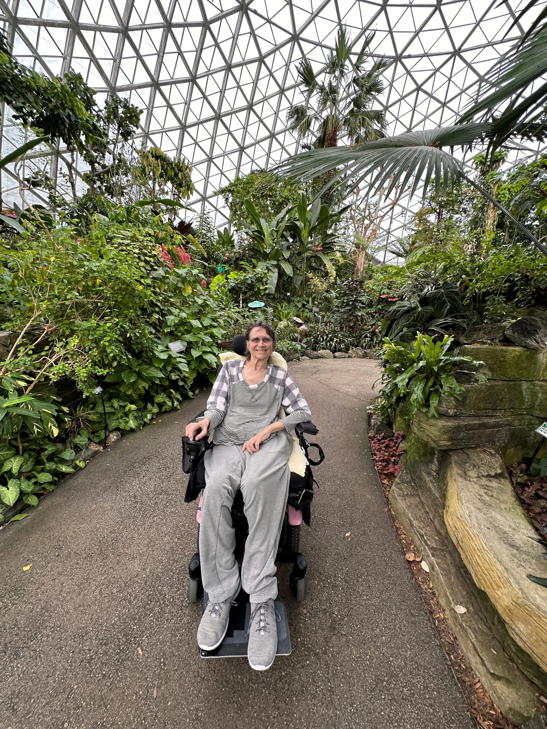 Smiling woman in a wheelchair on a paved path inside a glass-domed conservatory, surrounded by tropical plants.