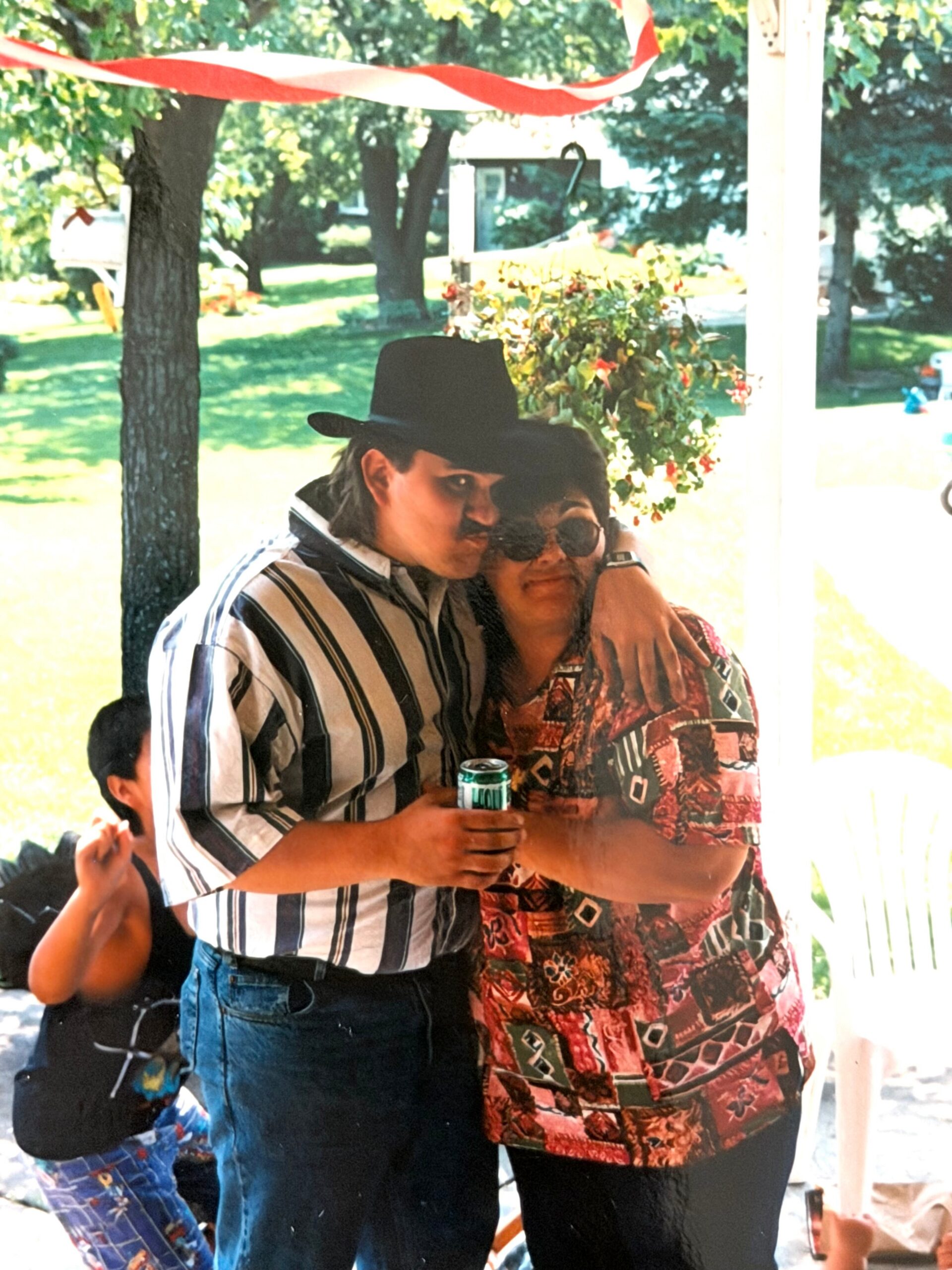 Couple hugging at an outdoor celebration; man in a striped shirt and black hat holds a can.