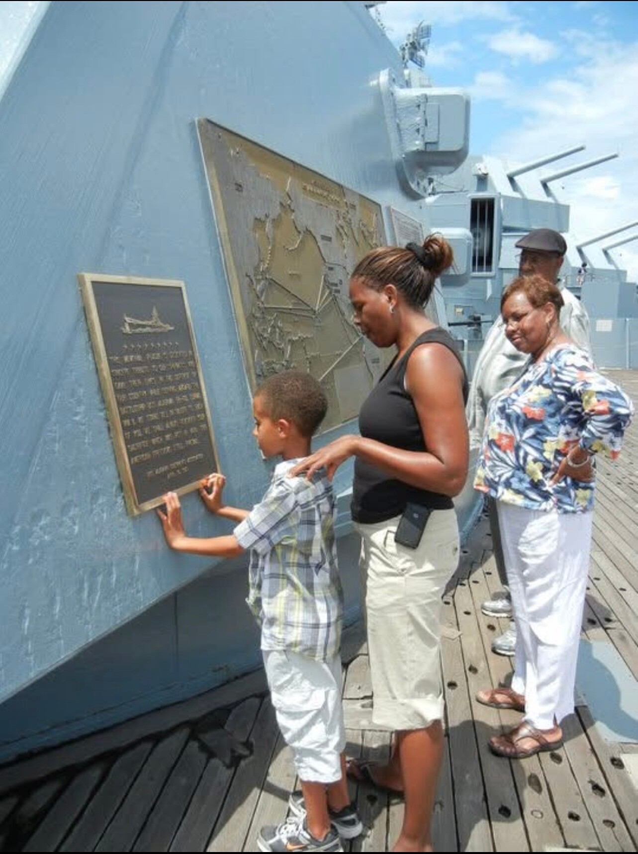 Family reads a commemorative plaque on the side of a blue naval ship, with a large map panel nearby.