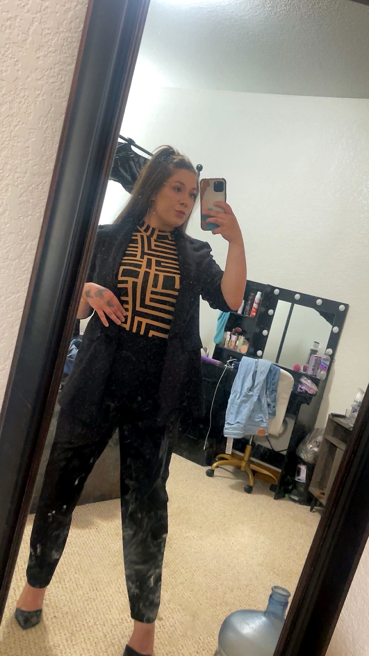 Self-portrait in a bedroom mirror: person wearing a black blazer over a geometric beige top and black pants, holding a phone to take a selfie.