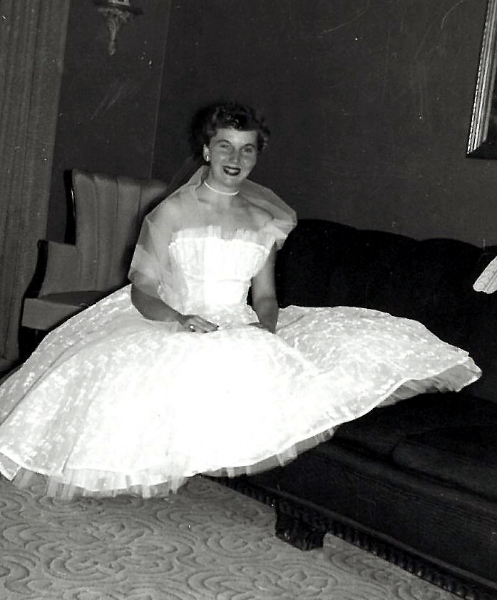 Black-and-white photo of a smiling bride in a full wedding gown seated on a sofa indoors, mid-20th century styling.
