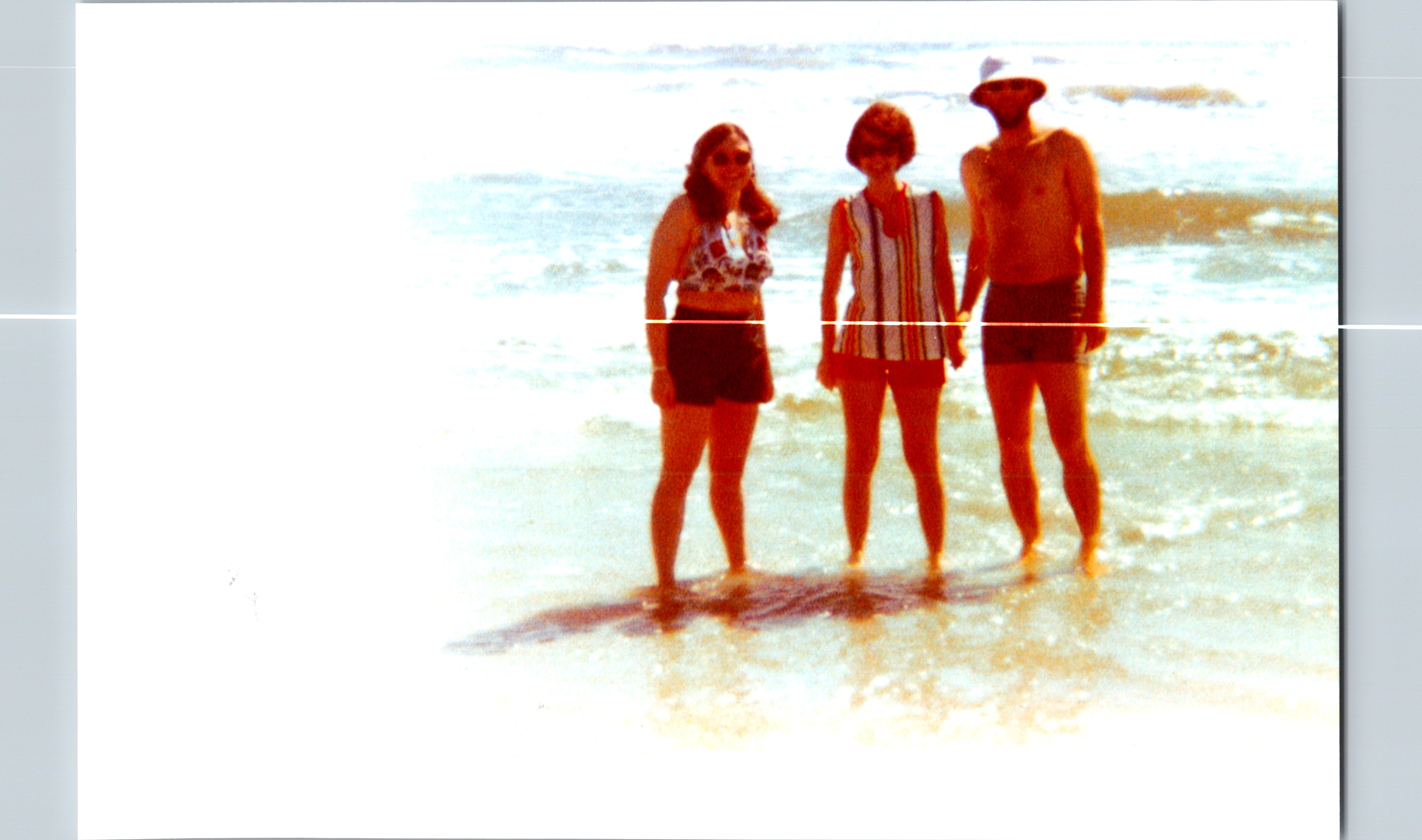 Three people in swimsuits stand at the shore with waves in the background, posing for a photo on the beach.