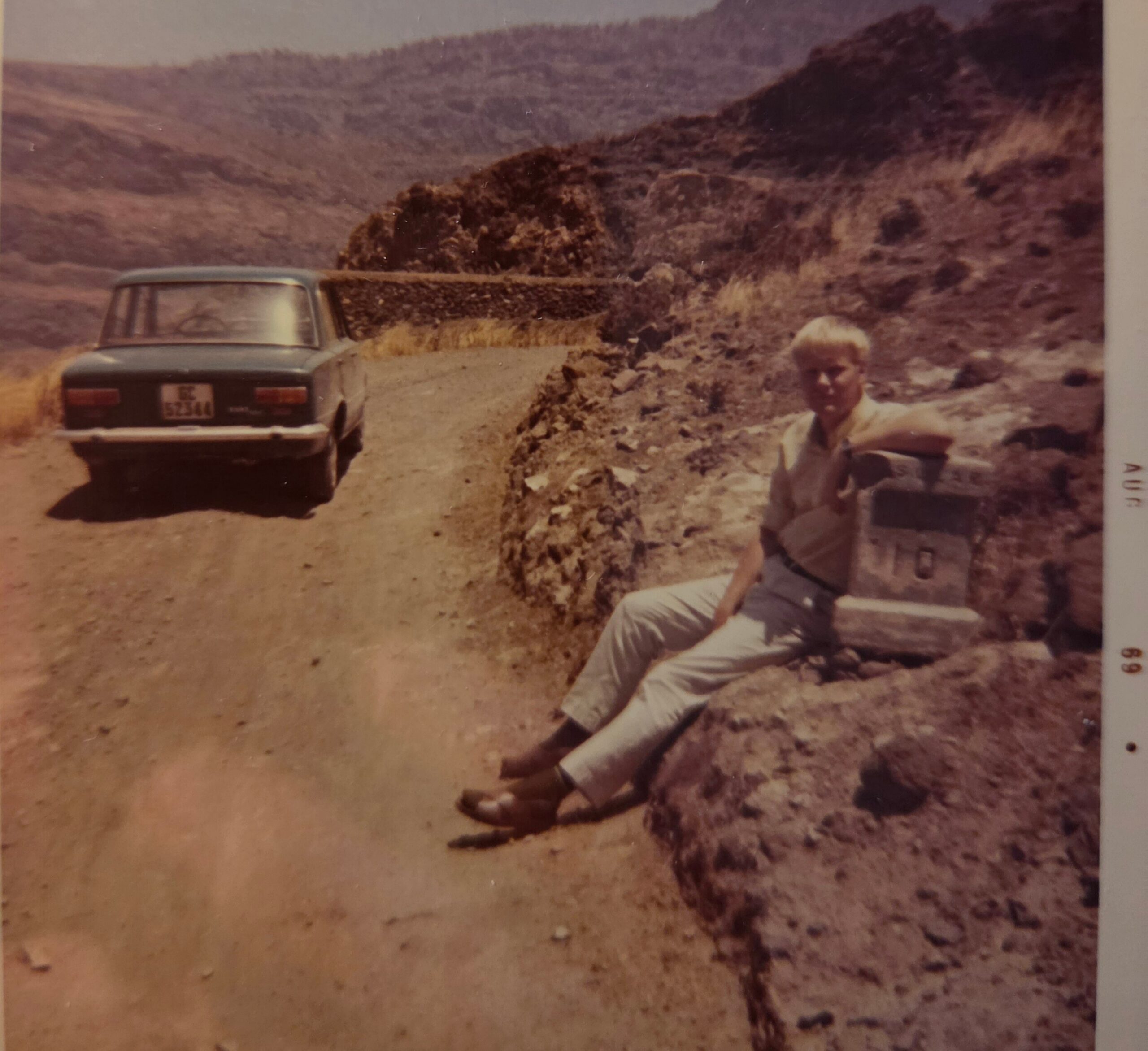 Dusty desert road scene with a vintage car parked on the left and a man sitting on rocks in the foreground, rocky hills in the background.