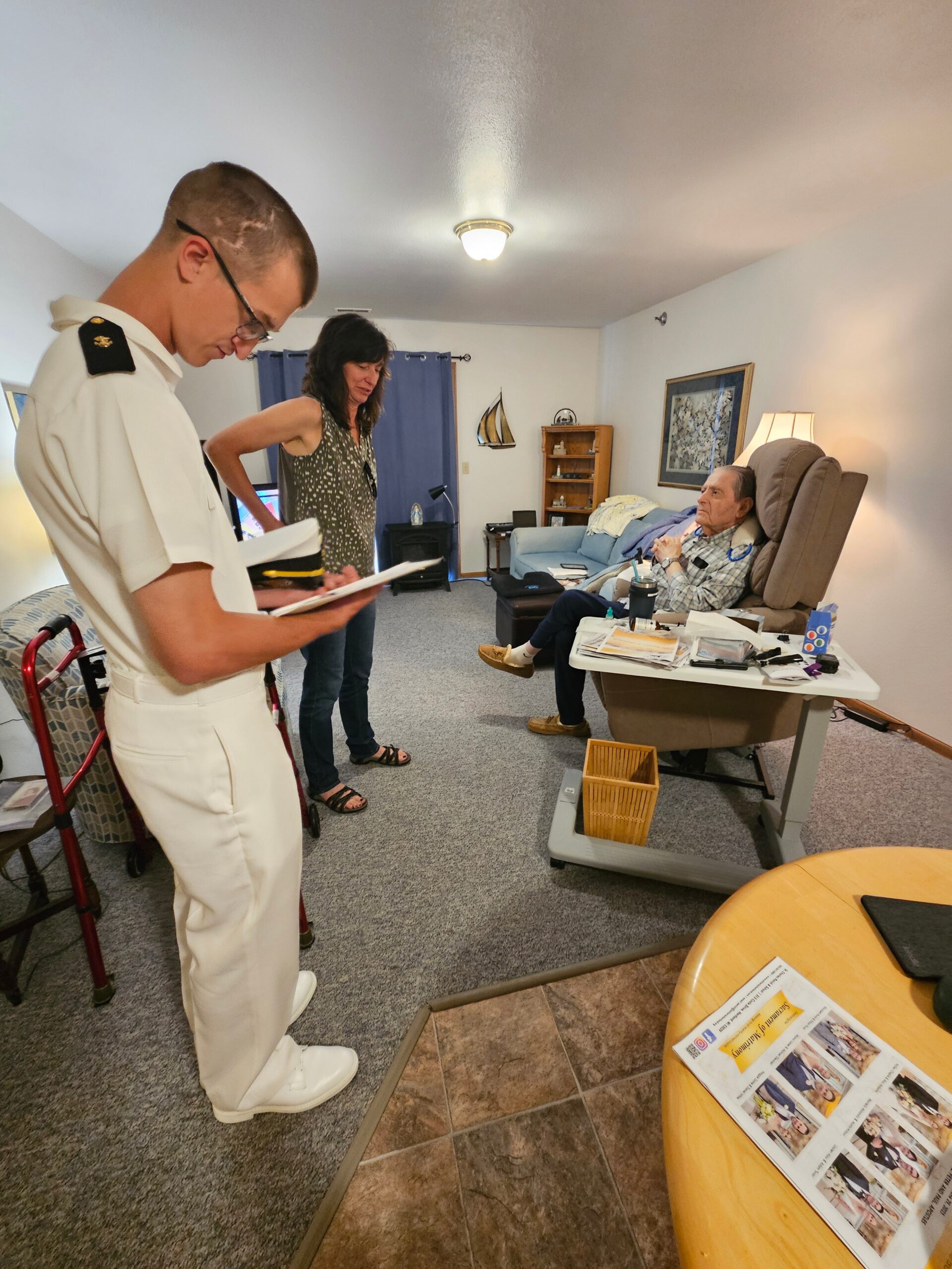 Elderly man sits in a recliner as a nurse in a white uniform and a woman review papers beside him in a cozy living room.