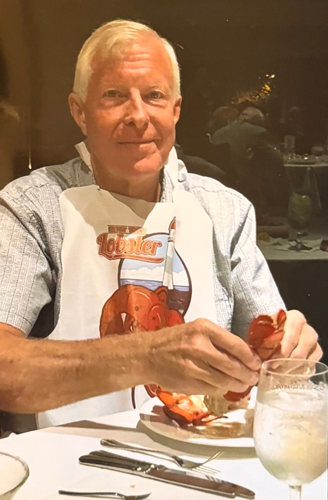 Smiling older man at a restaurant wearing a lobster-themed bib and cracking a lobster over a plate.