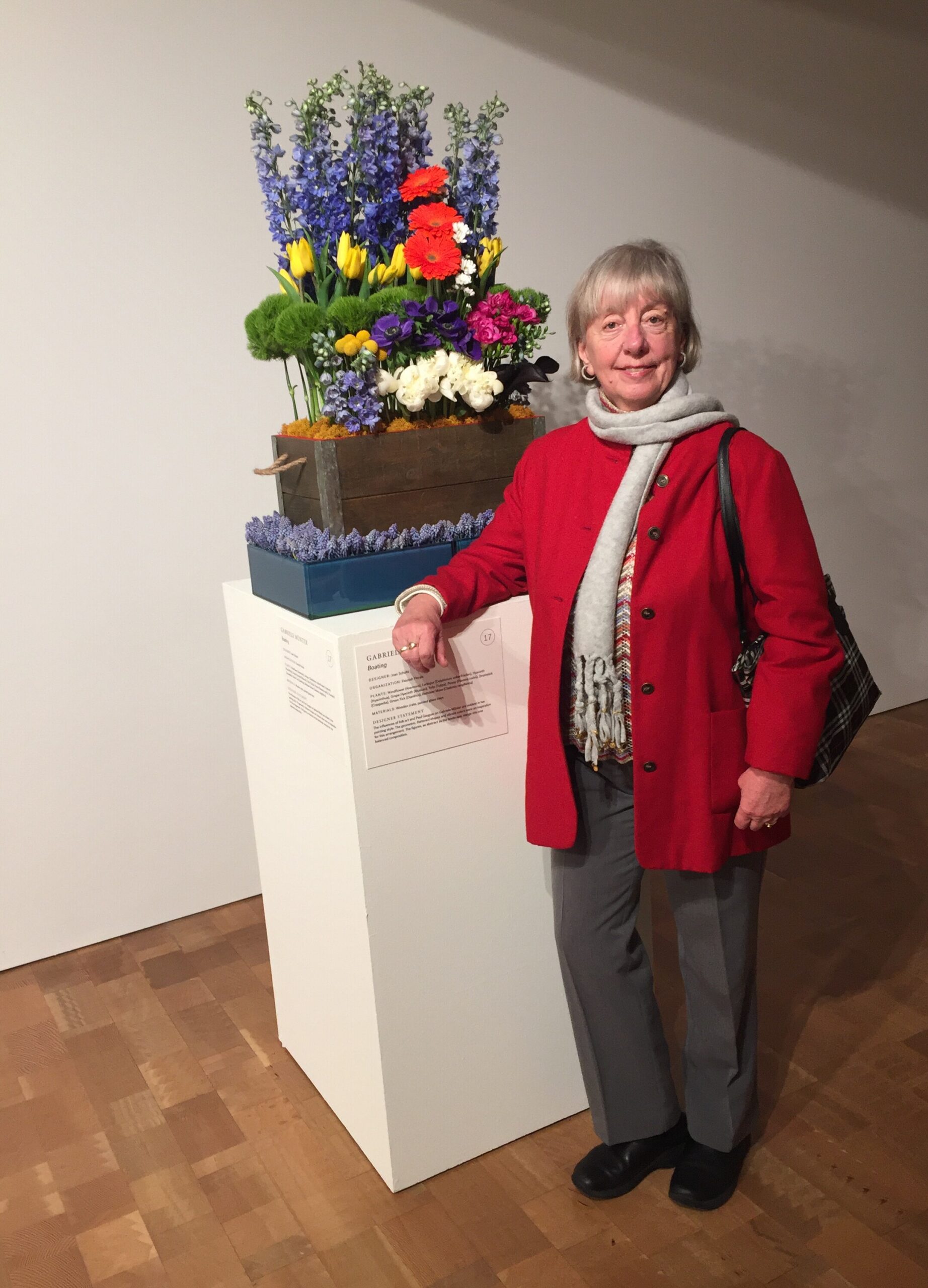 Smiling elderly woman in a red coat standing beside a tall multi-colored flower arrangement on a white pedestal in a gallery.