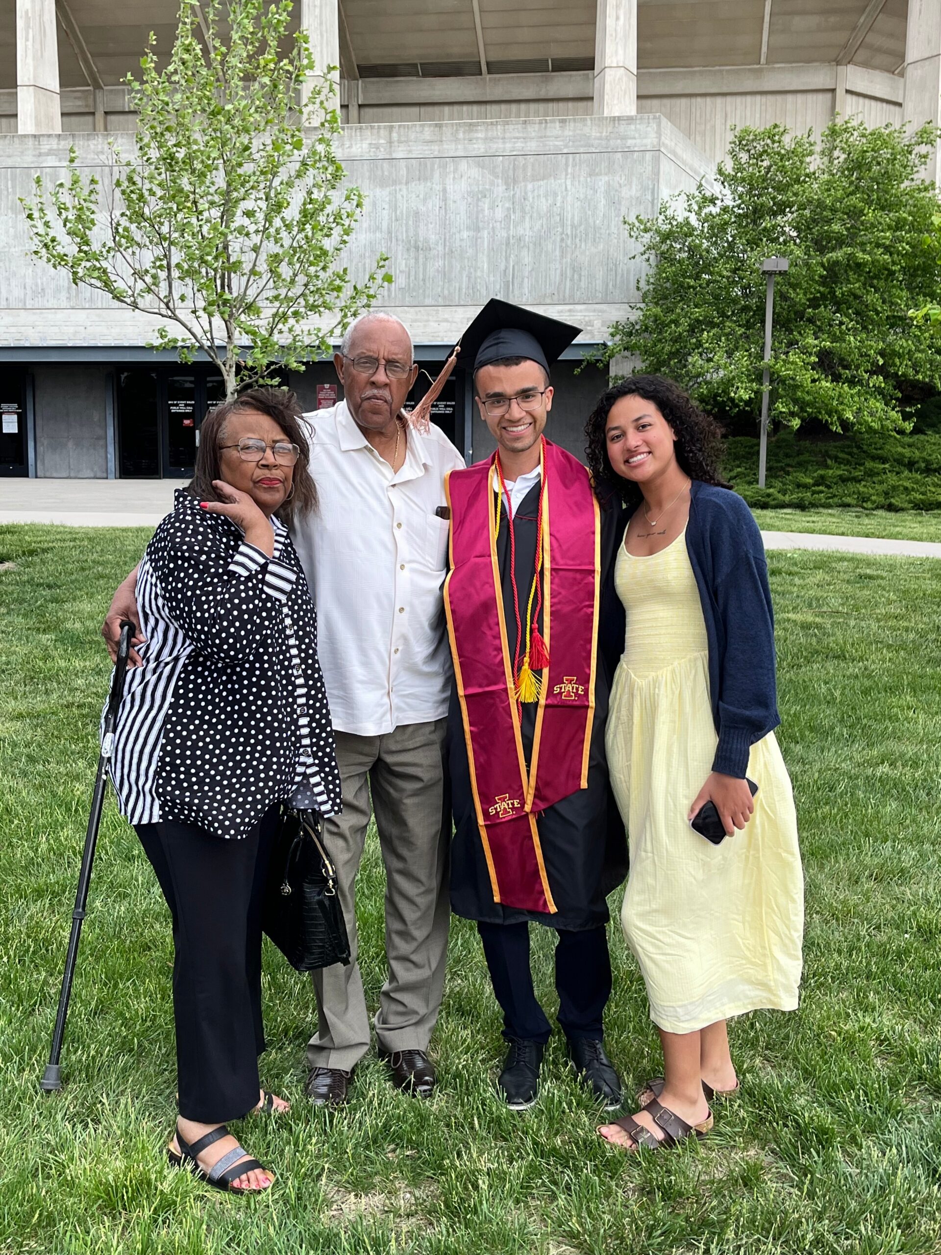 Graduate in cap and maroon gown poses with family on a grassy lawn in front of a concrete building.