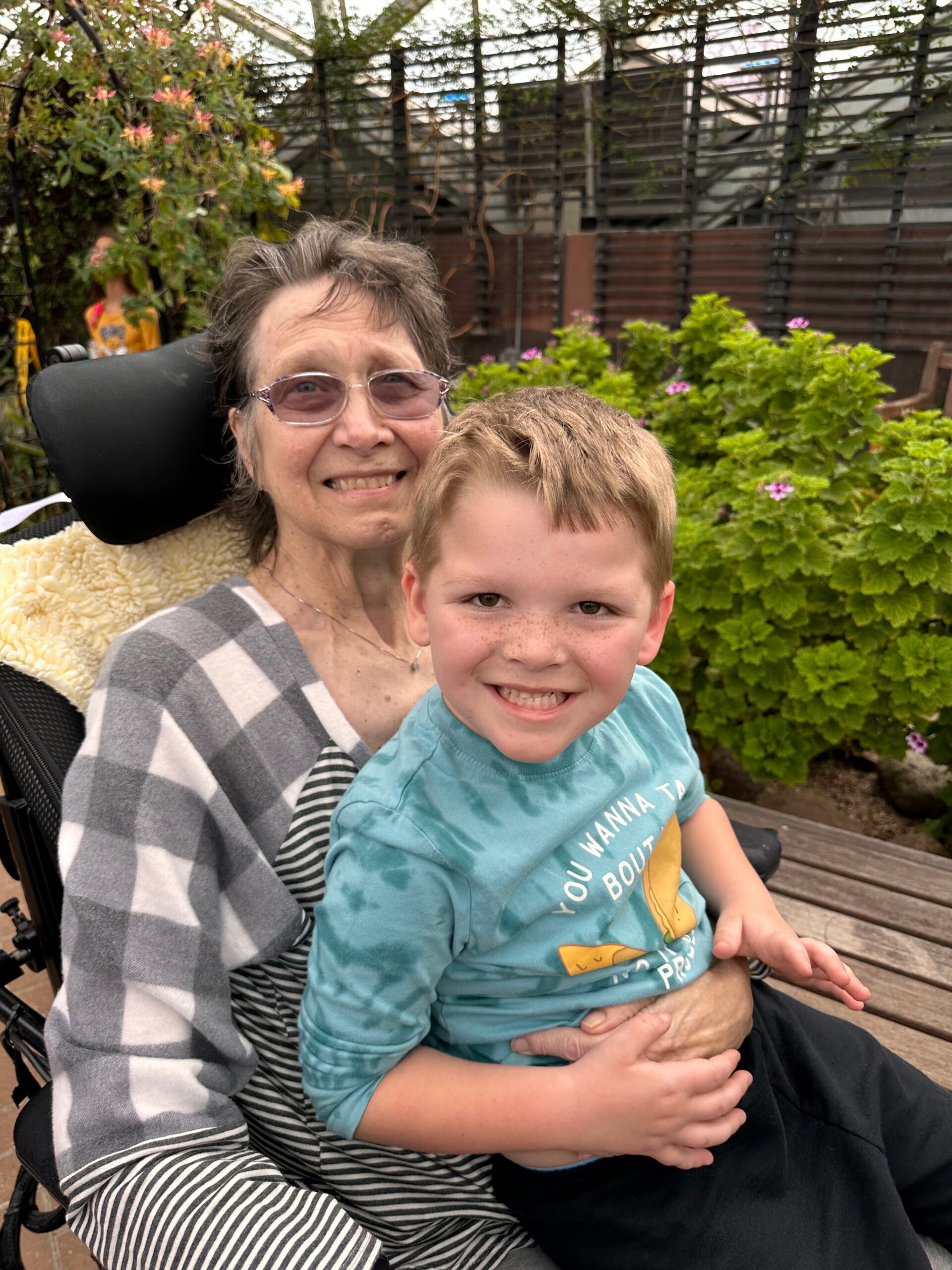 Smiling elderly woman in a wheelchair holding a young boy on her lap in a garden setting with green bushes behind them.