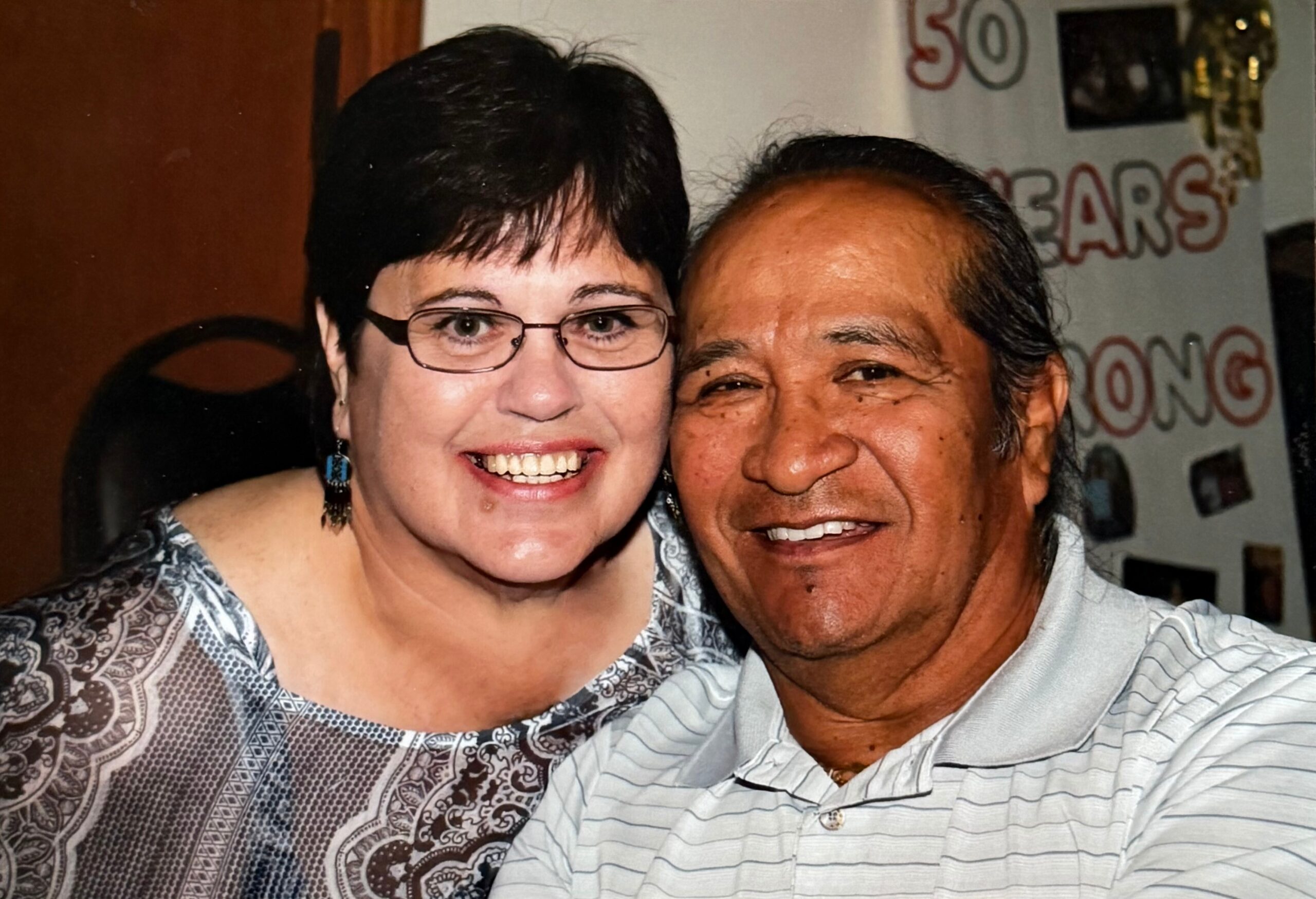 Smiling woman with short black hair and glasses beside a smiling man in a light-striped polo shirt indoors, posing for a selfie.