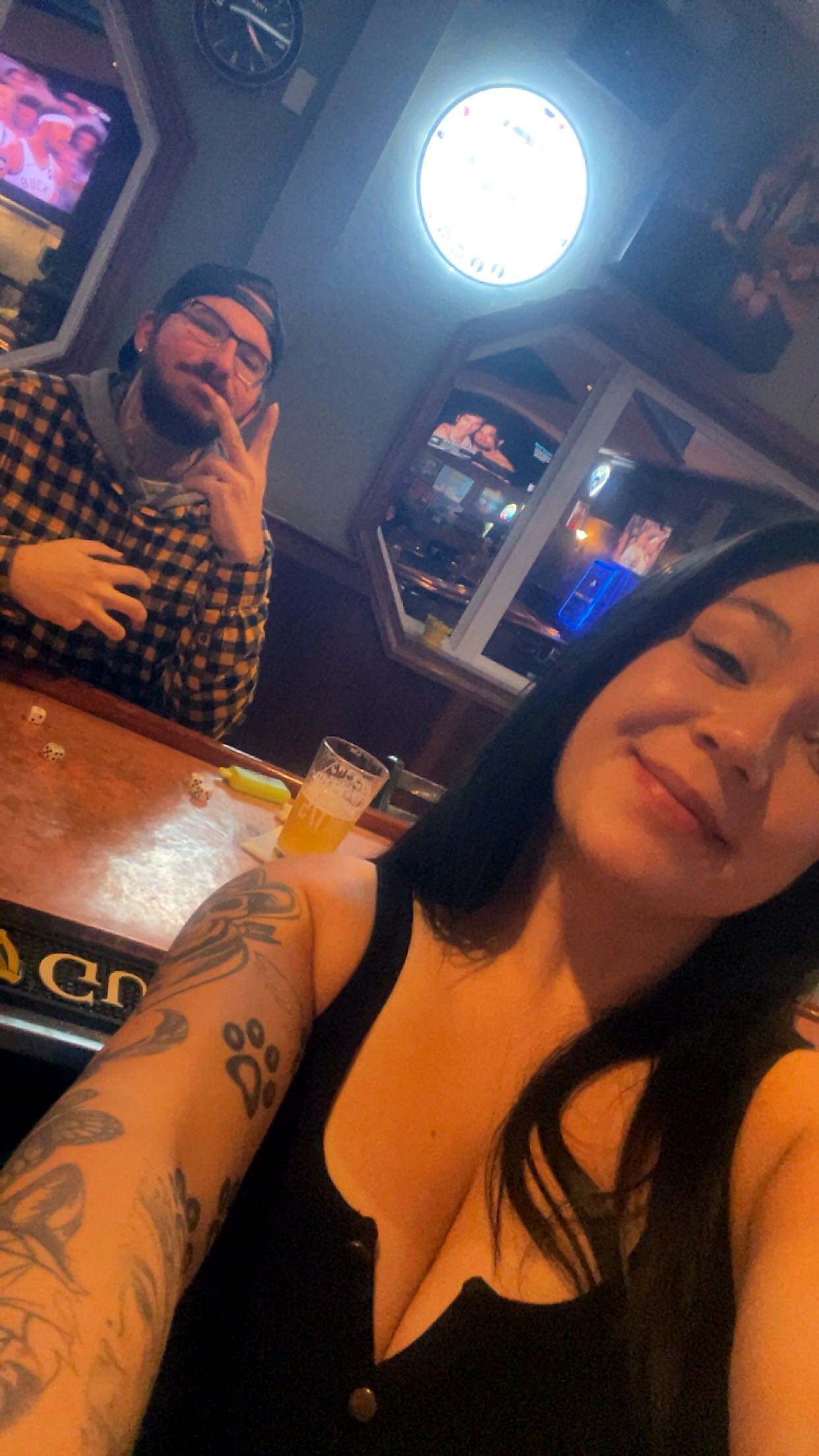 Two friends posing for a selfie at a bar, woman in foreground smiling with visible tattoos and a man in the background making a peace sign near a glass of beer on the counter