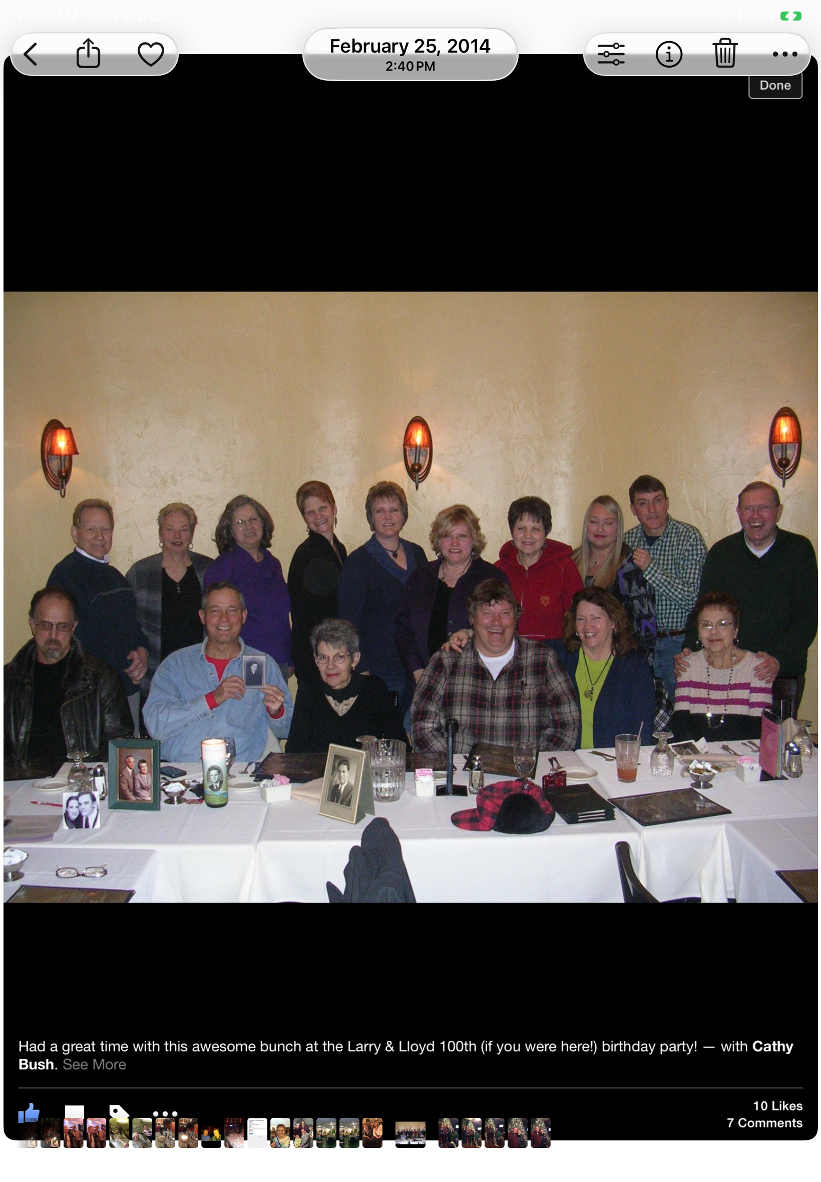 Group of about twenty people posing together at a birthday party around a long dining table with framed photos and candles on the table surface.