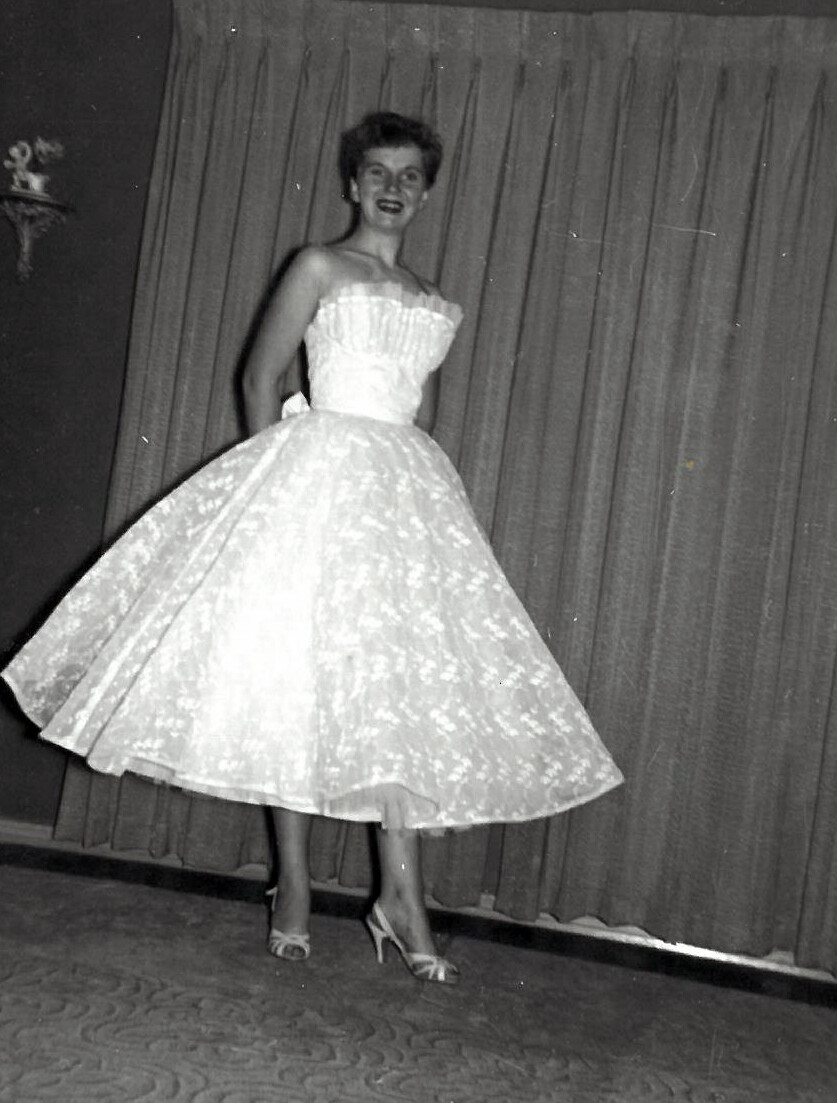 Woman in a strapless, full-skirted gown twirls on a stage, smiling, with curtains in the background.