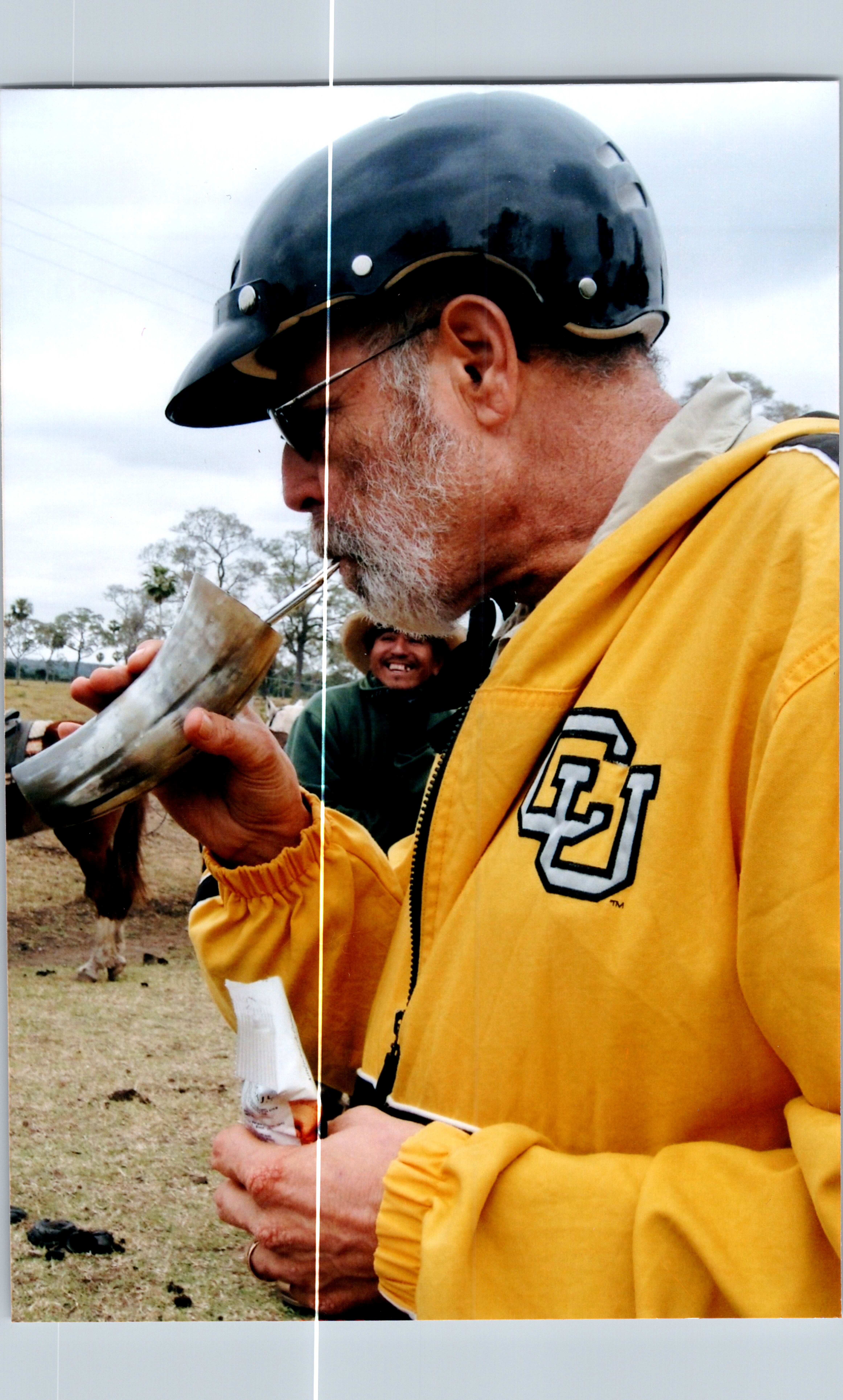 Man with a gray beard wearing a yellow jacket and black helmet, drinking from a metal horn outdoors while people and trees are visible in the background.