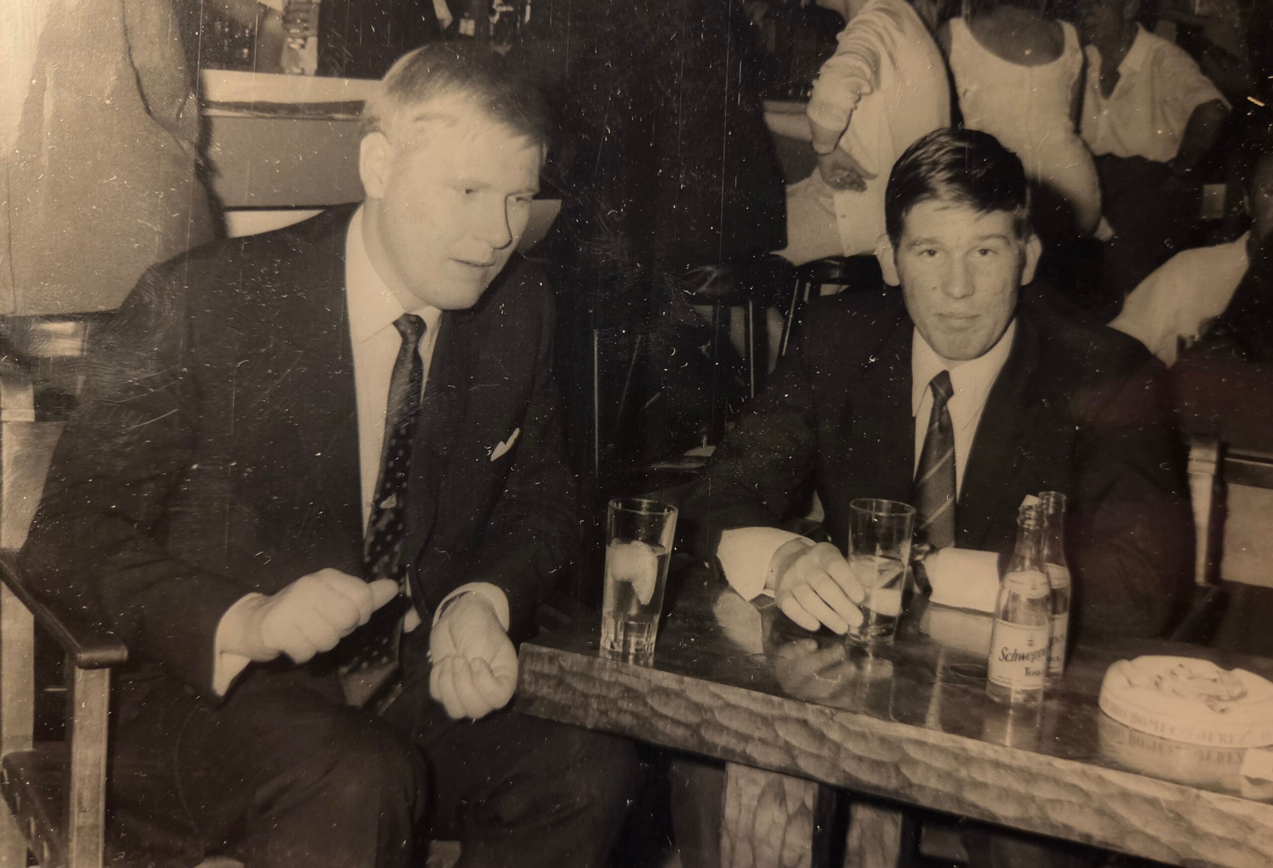 Two men in suits sit at a table with drinks during a formal gathering, posing for a photo in a black-and-white scene of the event, 1960s era likely.