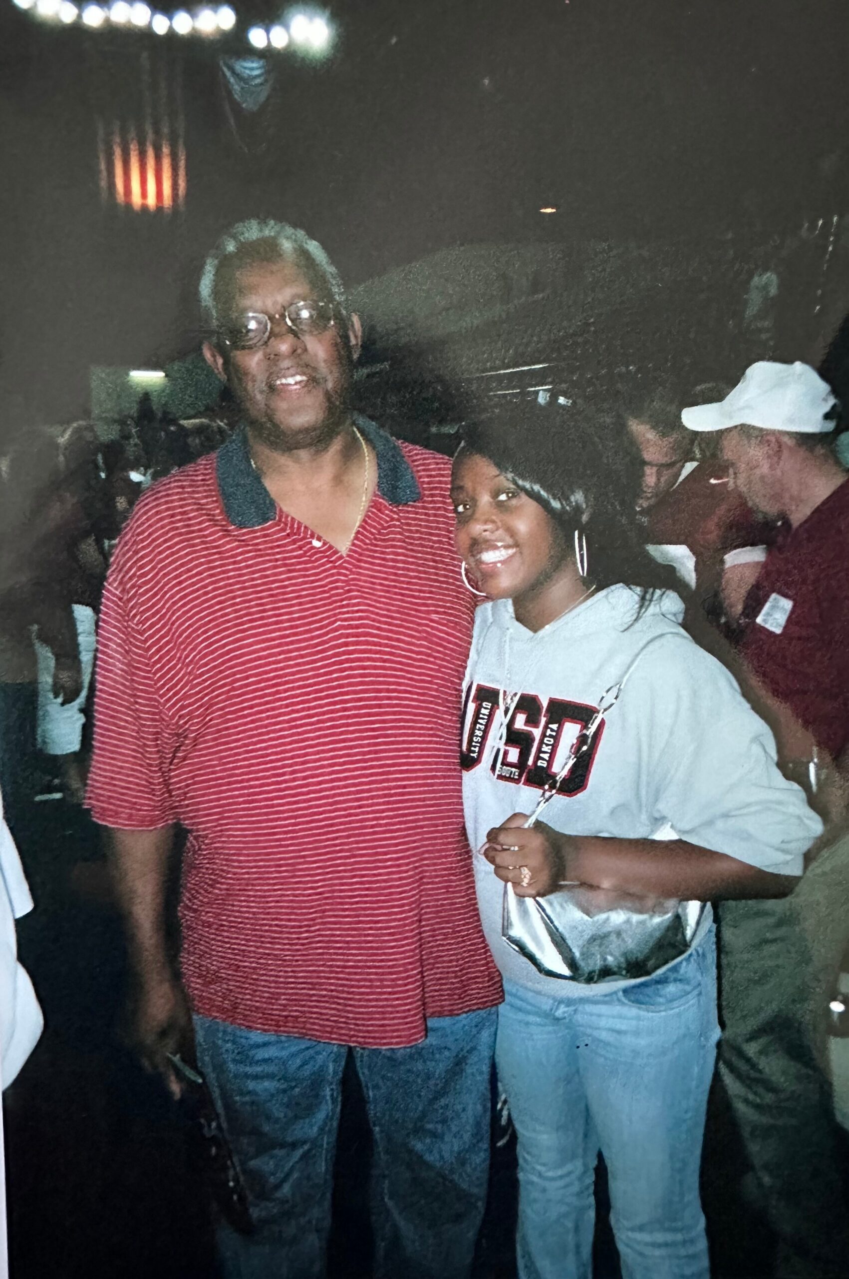 Two people pose together at a nighttime event: a man in a red striped polo shirt with glasses and a smile, standing beside a woman in a gray hoodie with a lanyard, both looking at the camera.