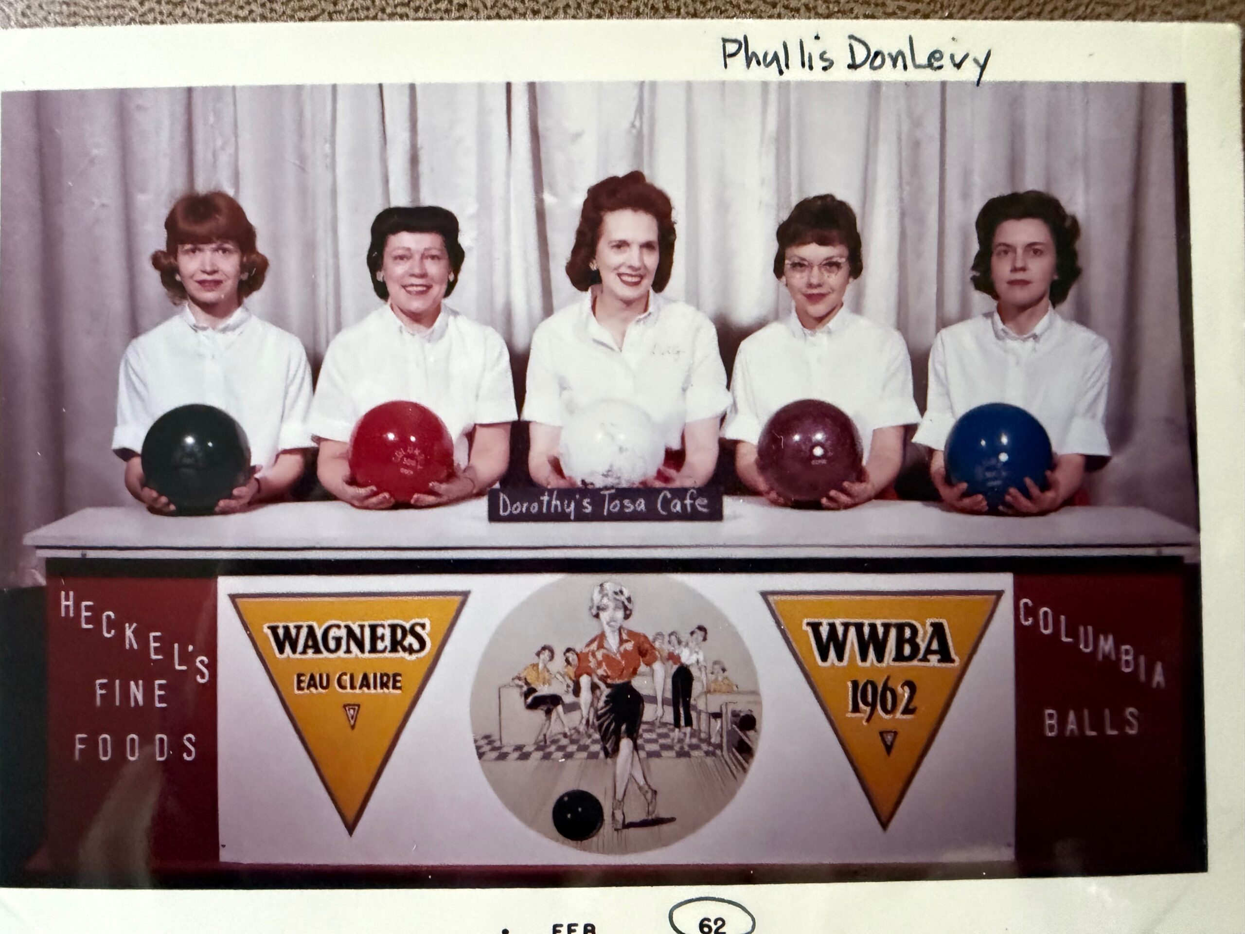 Five women in white uniforms pose with bowling balls in front of a vintage cafe sign and retro posters.