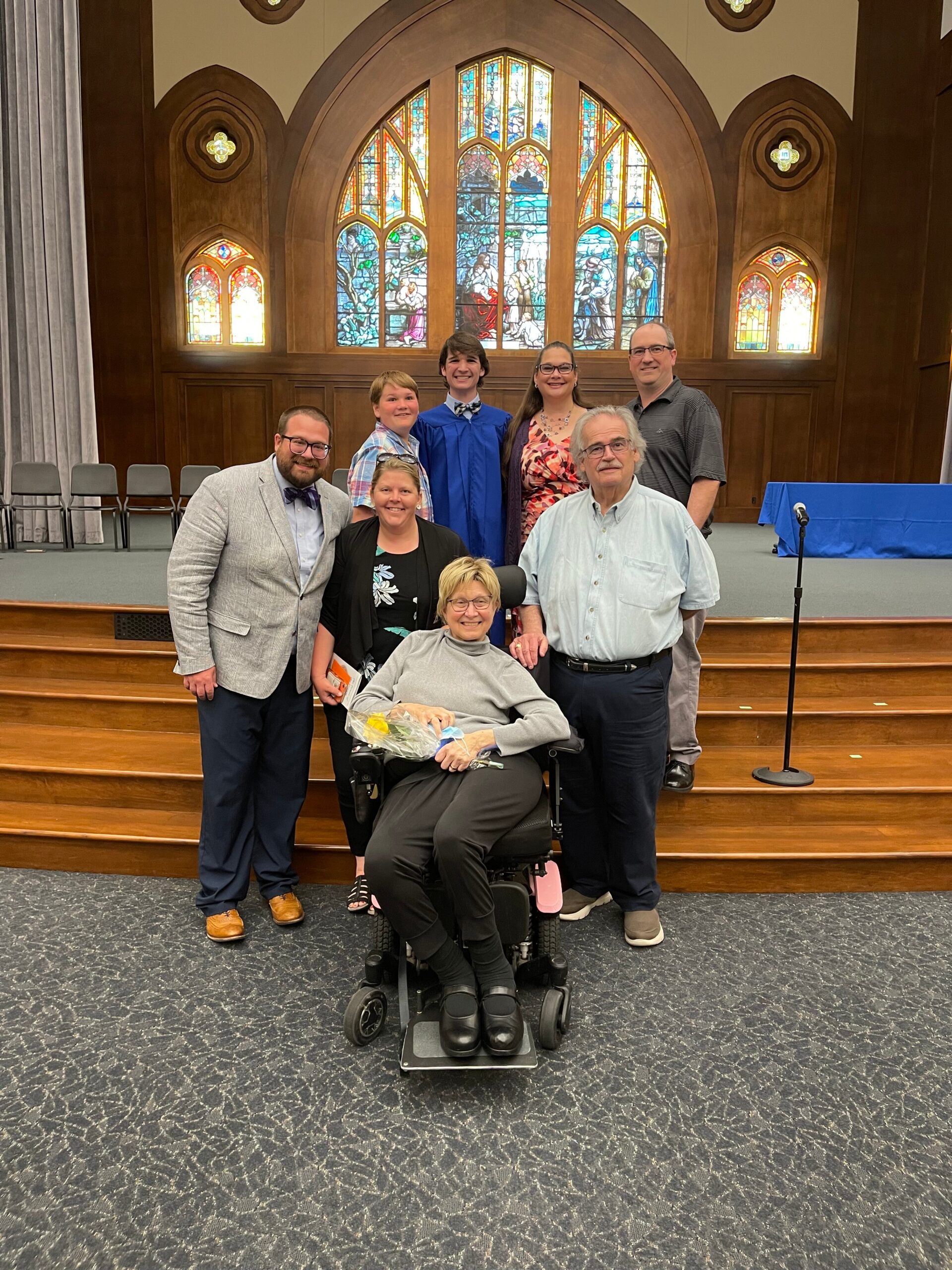 Group of eight people posing on church steps, with a woman in a wheelchair at the front, in front of a large stained-glass window.