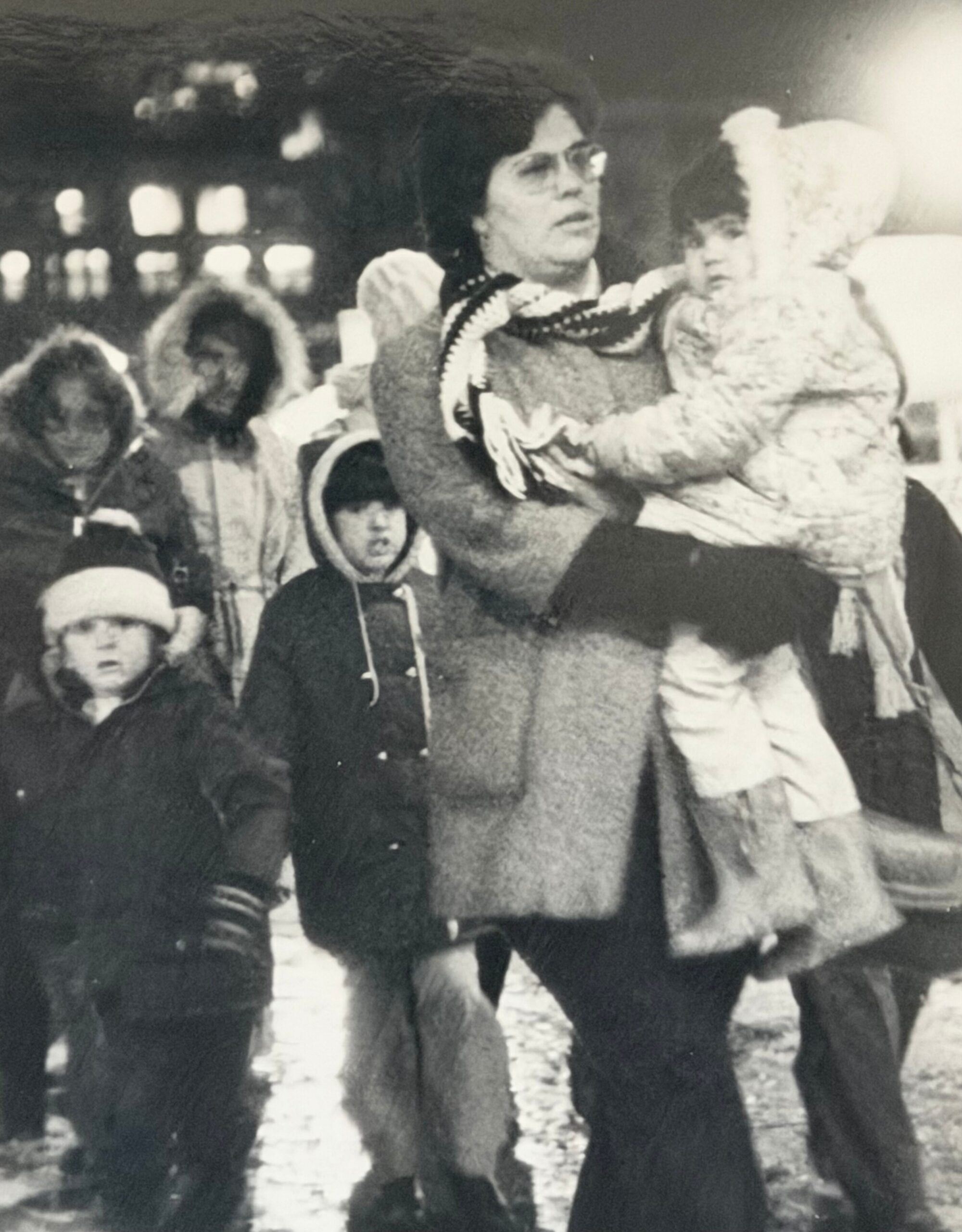 Woman carrying a child while surrounded by people in winter clothing at an indoor gathering (black-and-white).