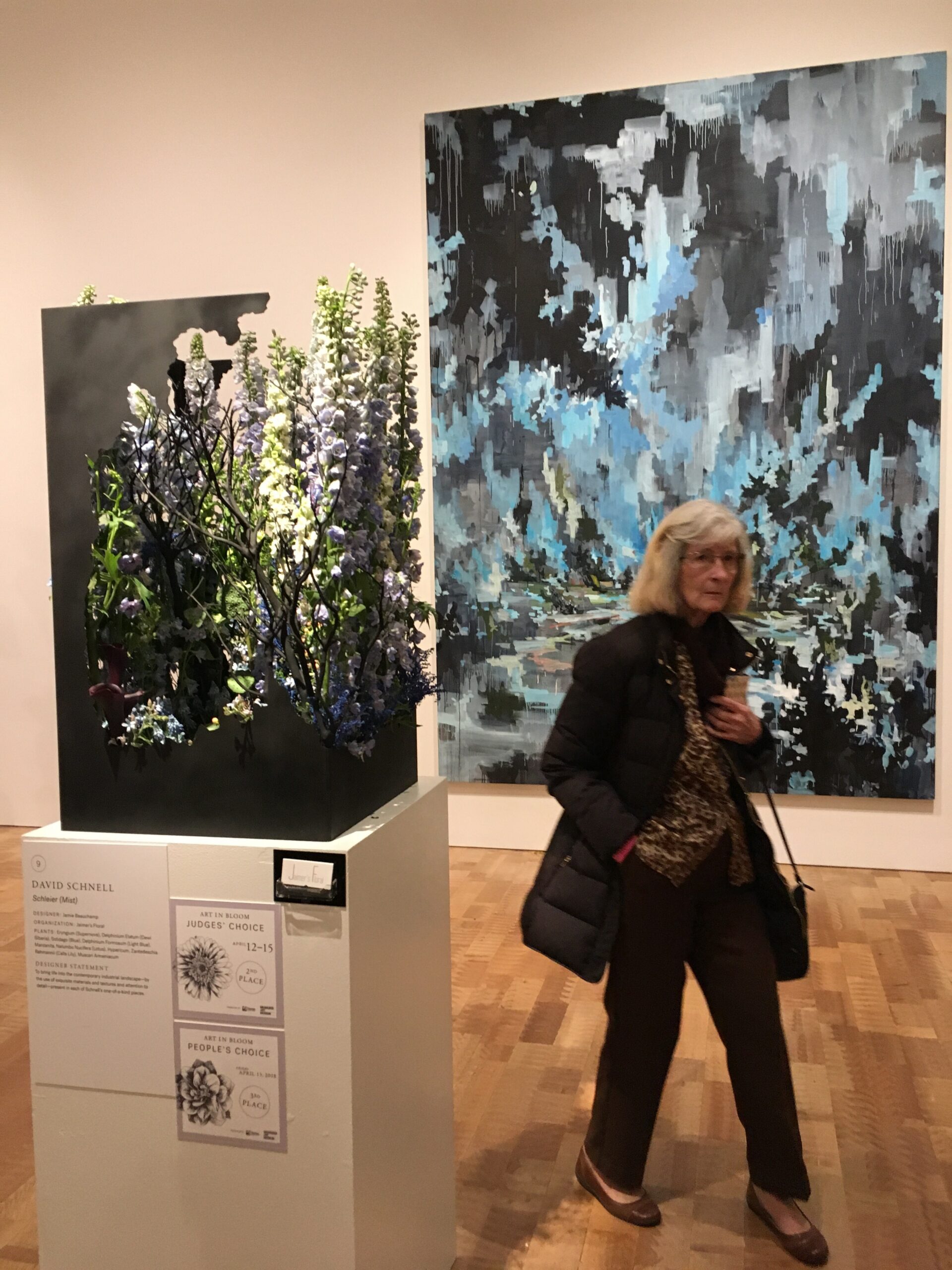 Large abstract blue-gray painting on the wall beside a black display case filled with tall flowering branches in purple, white, and green; a woman in dark clothing walks past in the foreground.