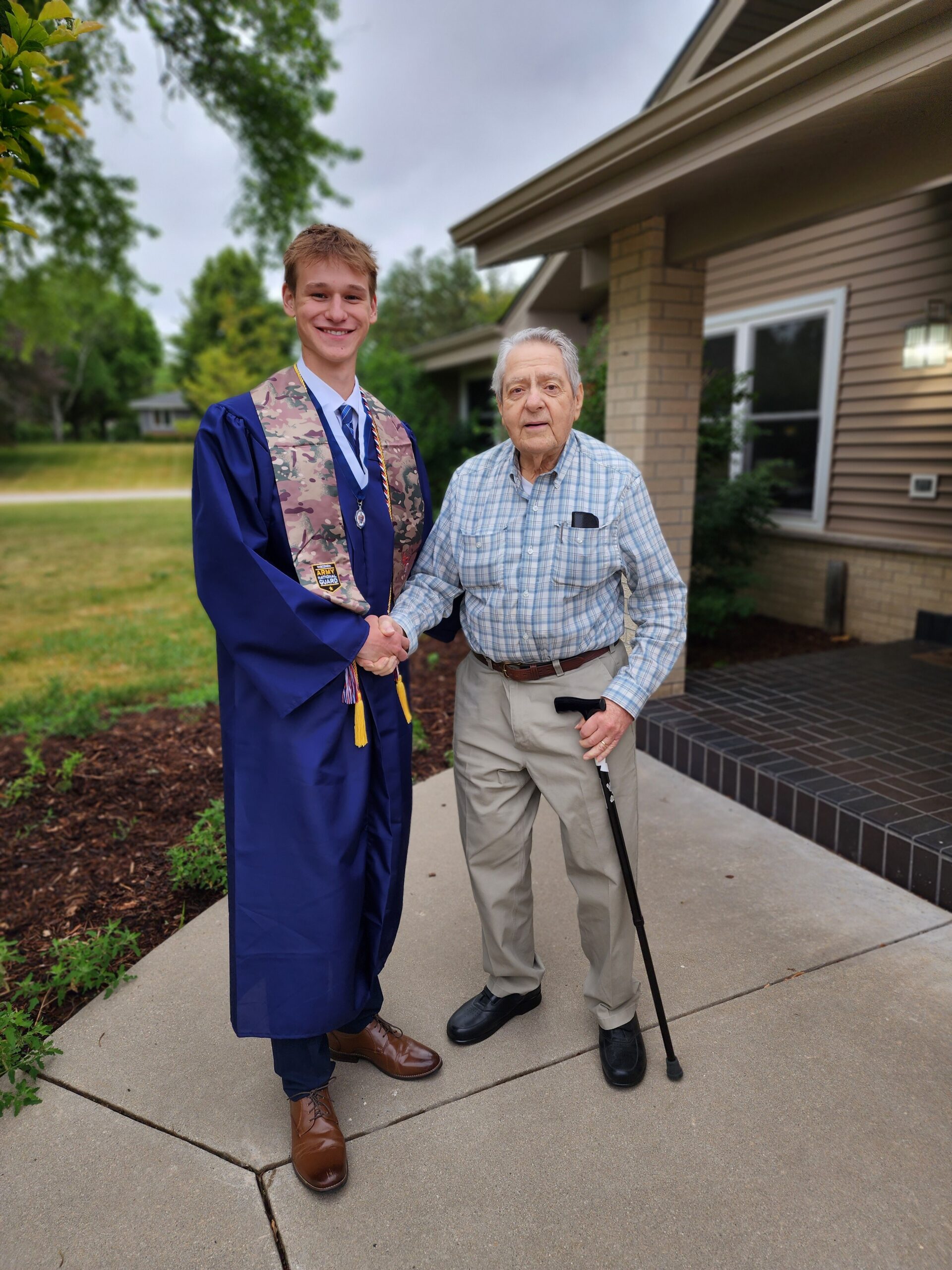 A smiling graduate in a blue cap and gown shakes hands with an elderly man on a driveway outside a house, both facing the camera.