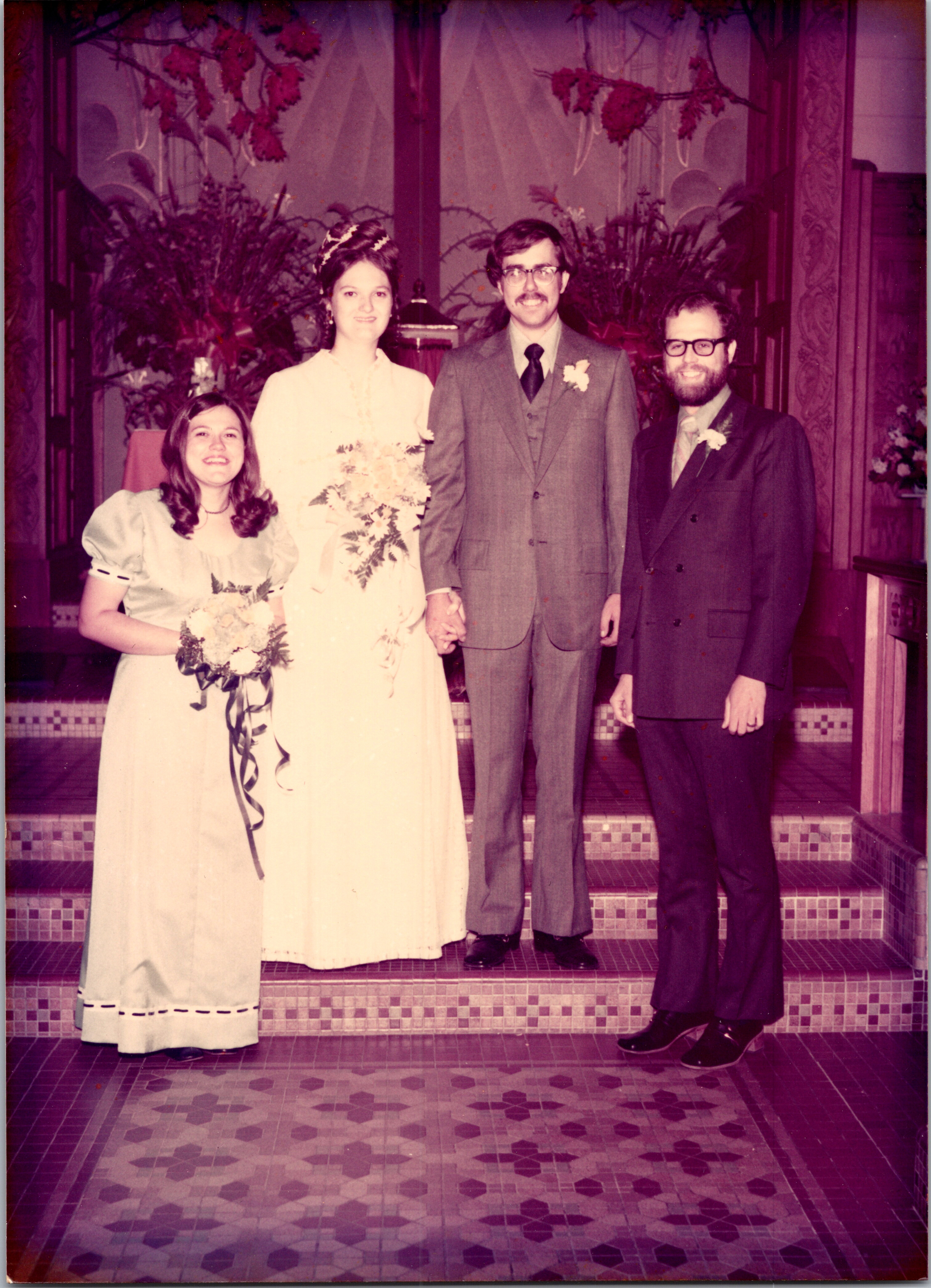 Four people pose for a wedding photo on tiled steps inside a decorated hall: from left, bridesmaid in a light gown, bride in white holding a bouquet, and two men in suits (groom in gray, groomsman in darker suit).
